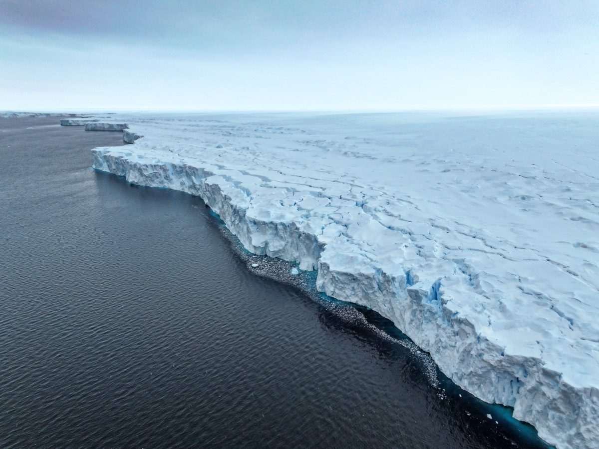 Aerial view of the edge of a large glacier, where it meets the ocean, with the view of the large pale ice mass including its serrated edge which is contiguous with a stony beach area, with the whole thing receding in size towards the horizon