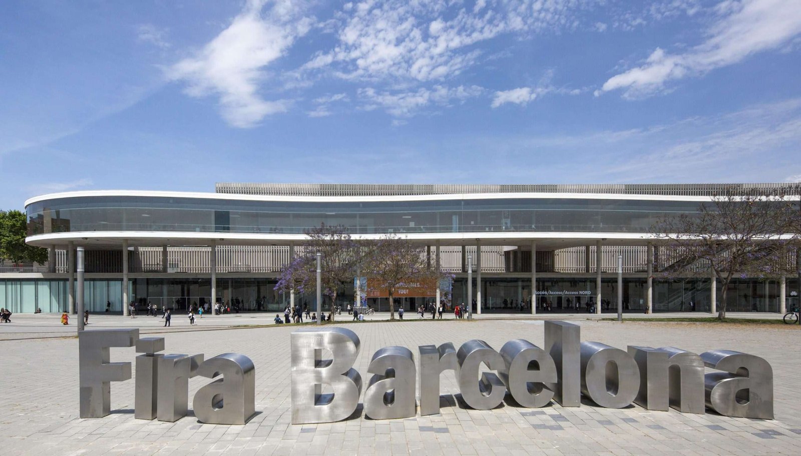 Exterior of large exhibition space with people visible as tiny figures near the foot of the large, low-lying, modern-looking structure, and in the foreground the letters of the words 'Fira Barcelona' have been realized as large, metallic scultpures resting on the concrete-paved ground in front of the building