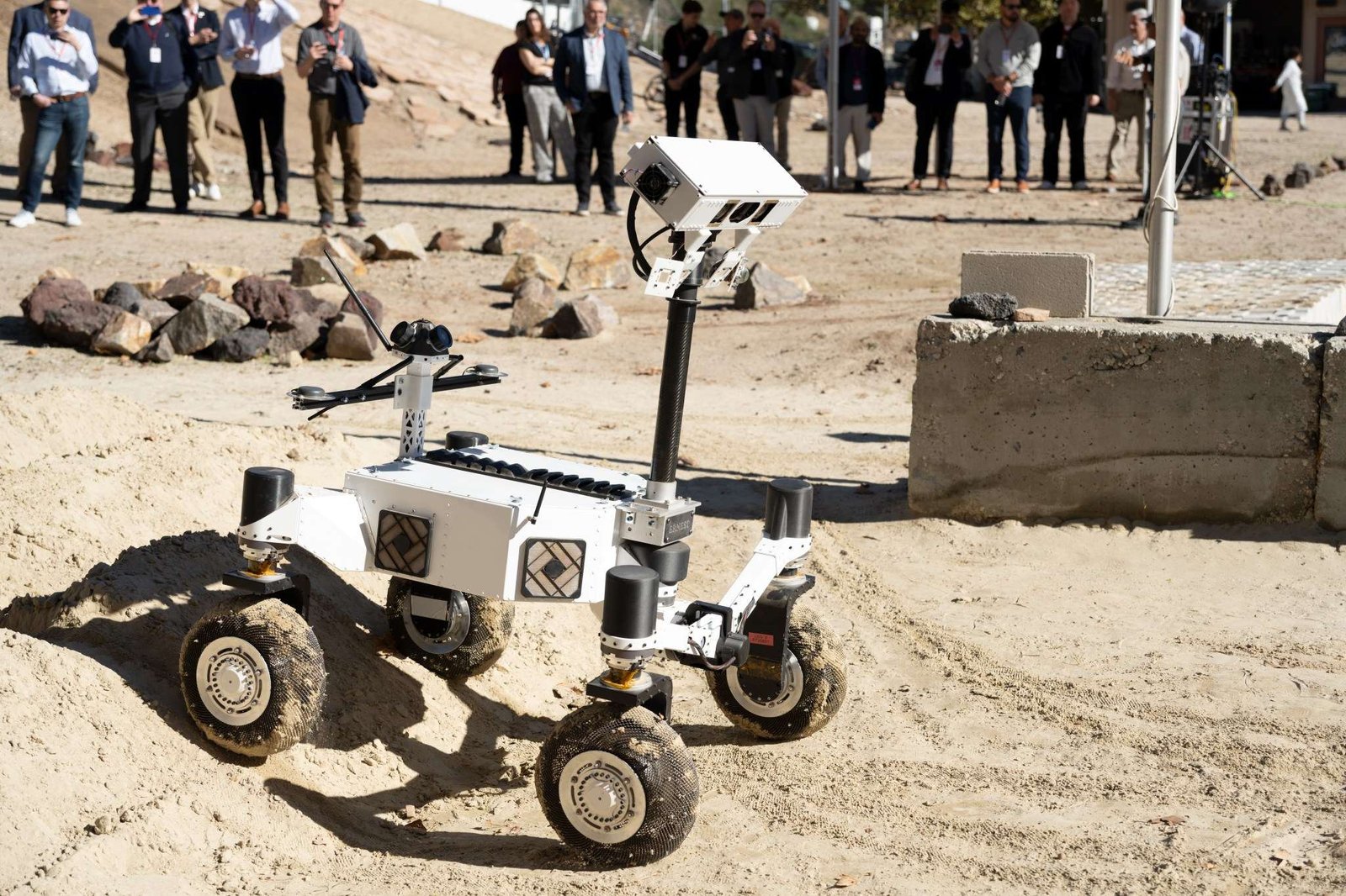 A rover drives down an incline as a group of people watch from a distance.