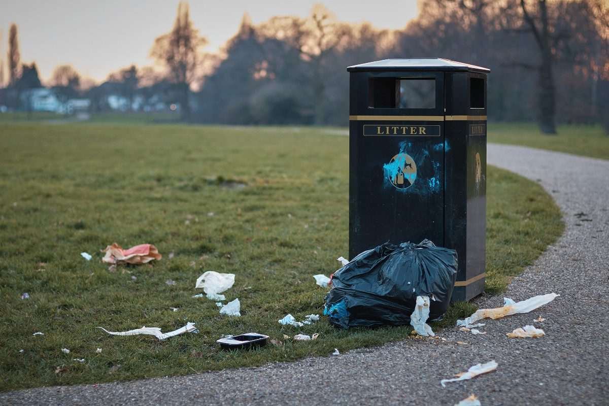 A black municipal litter bin in a public park with litter scattered on the grass in its vicinity