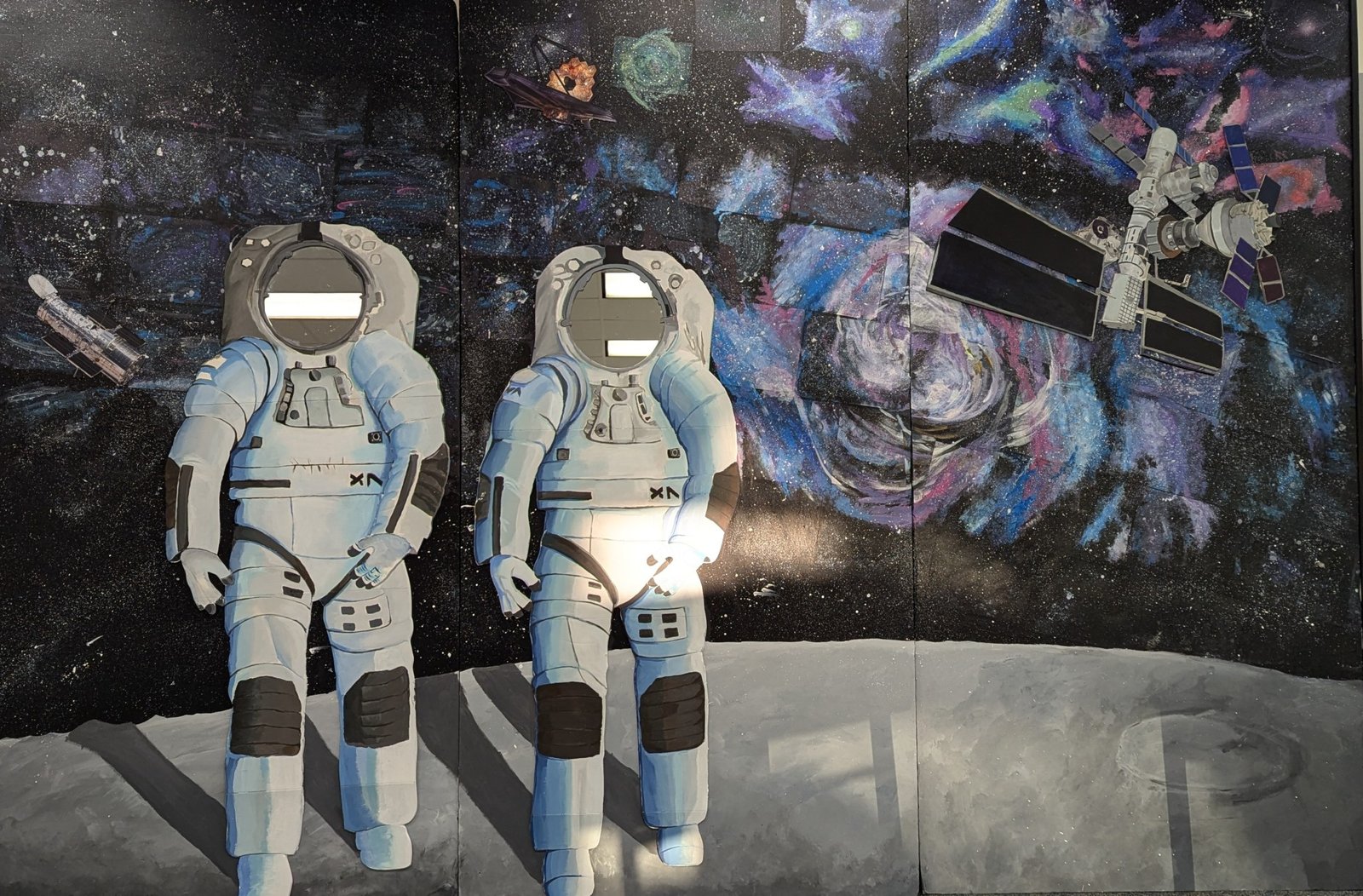 A mural showcases two spacesuit cutouts on a lunar surface, allowing visitors to pose as astronauts. The backdrop features a depiction of space, with colorful galaxies, the Hubble Space Telescope, and a satellite orbiting a distant planet.
