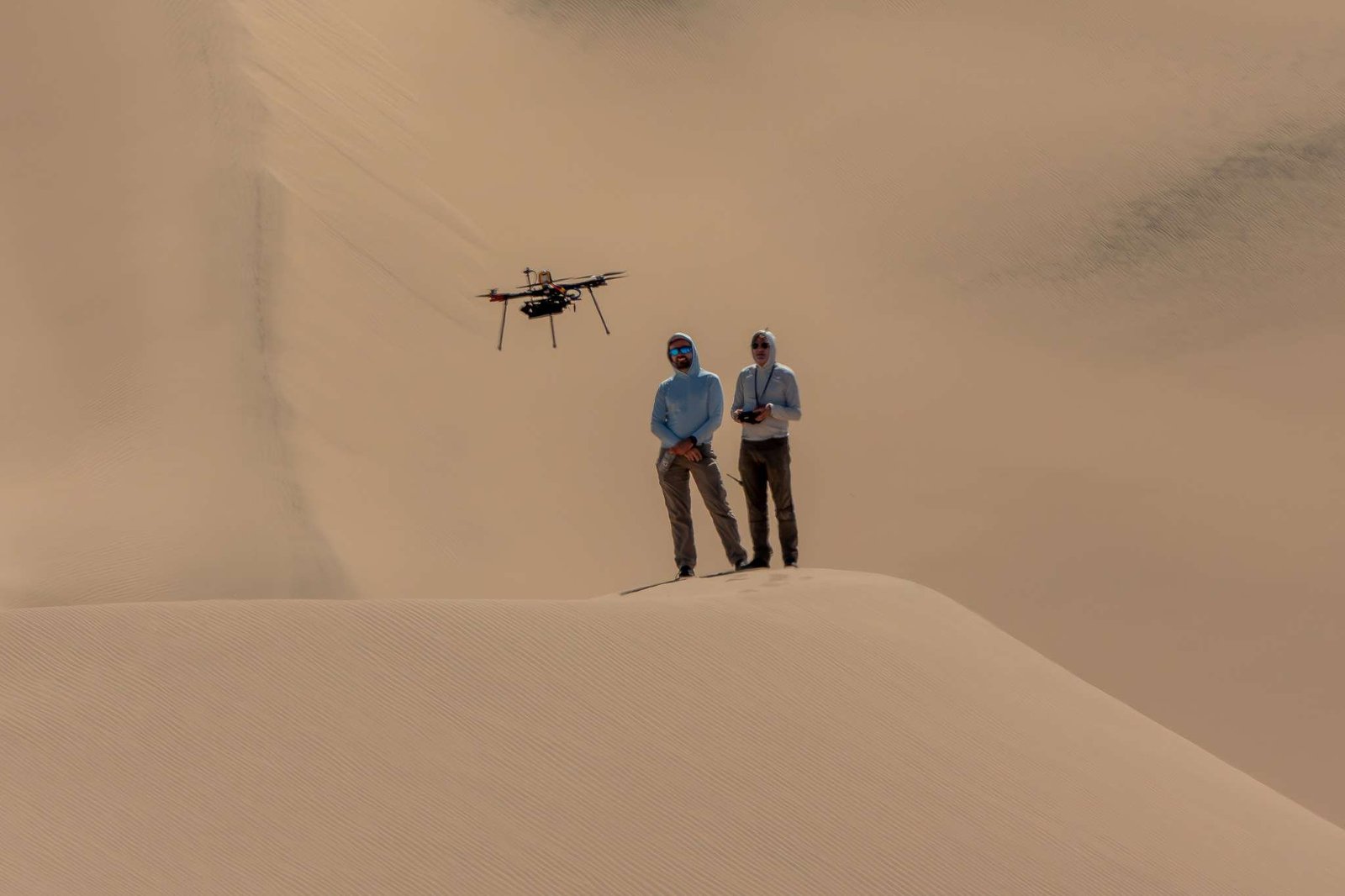 Two people stand atop a sand dune, facing the camera. They are both looking at a drone flying in front of them. The person on the right holds a controller. Other sand dunes behind them make up the background of the entire image.