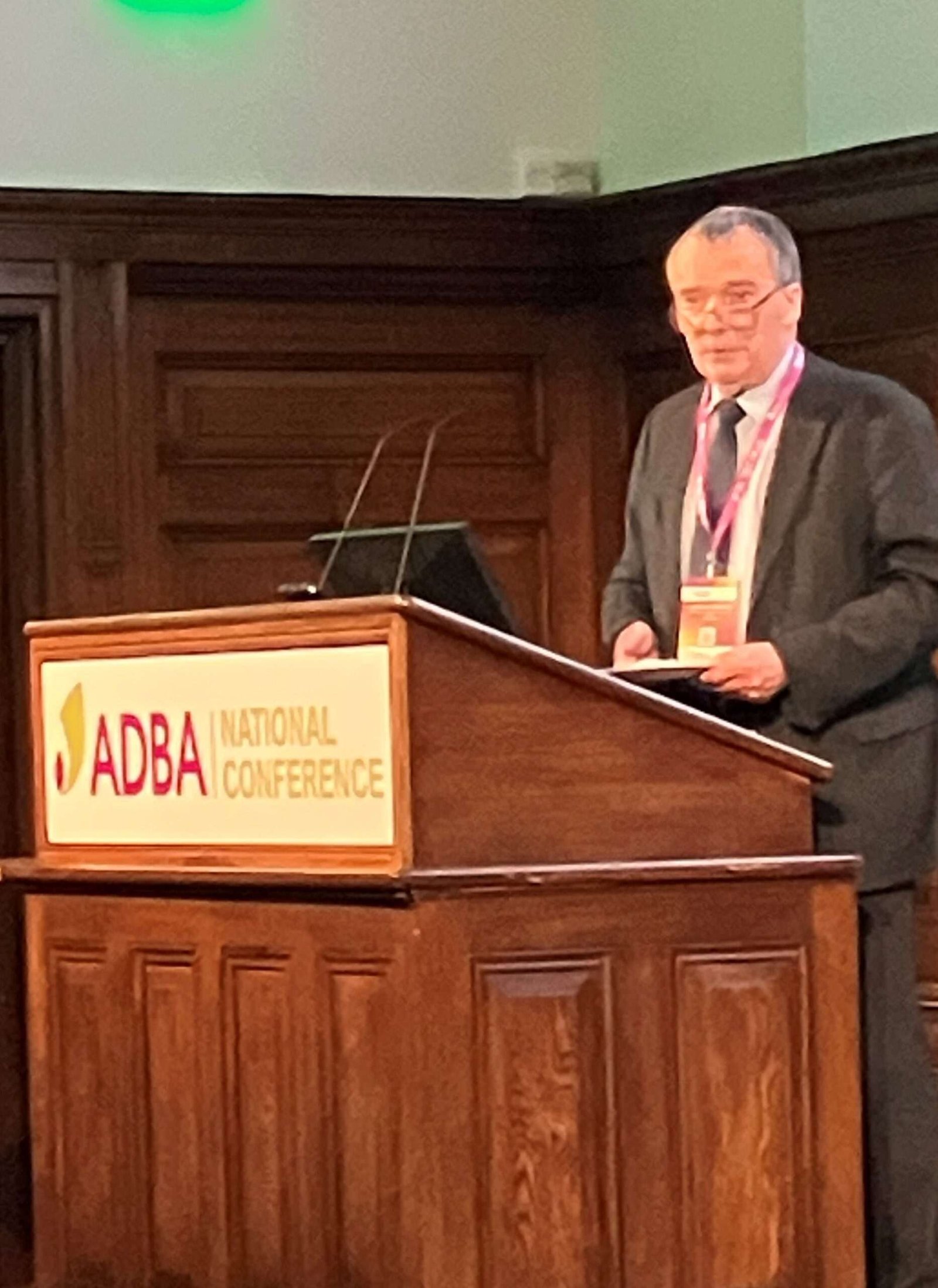 A middle-aged man in formal attire stands at a wooden lectern upon which is a coloured board with prominent wording 'ADBA National Conference'