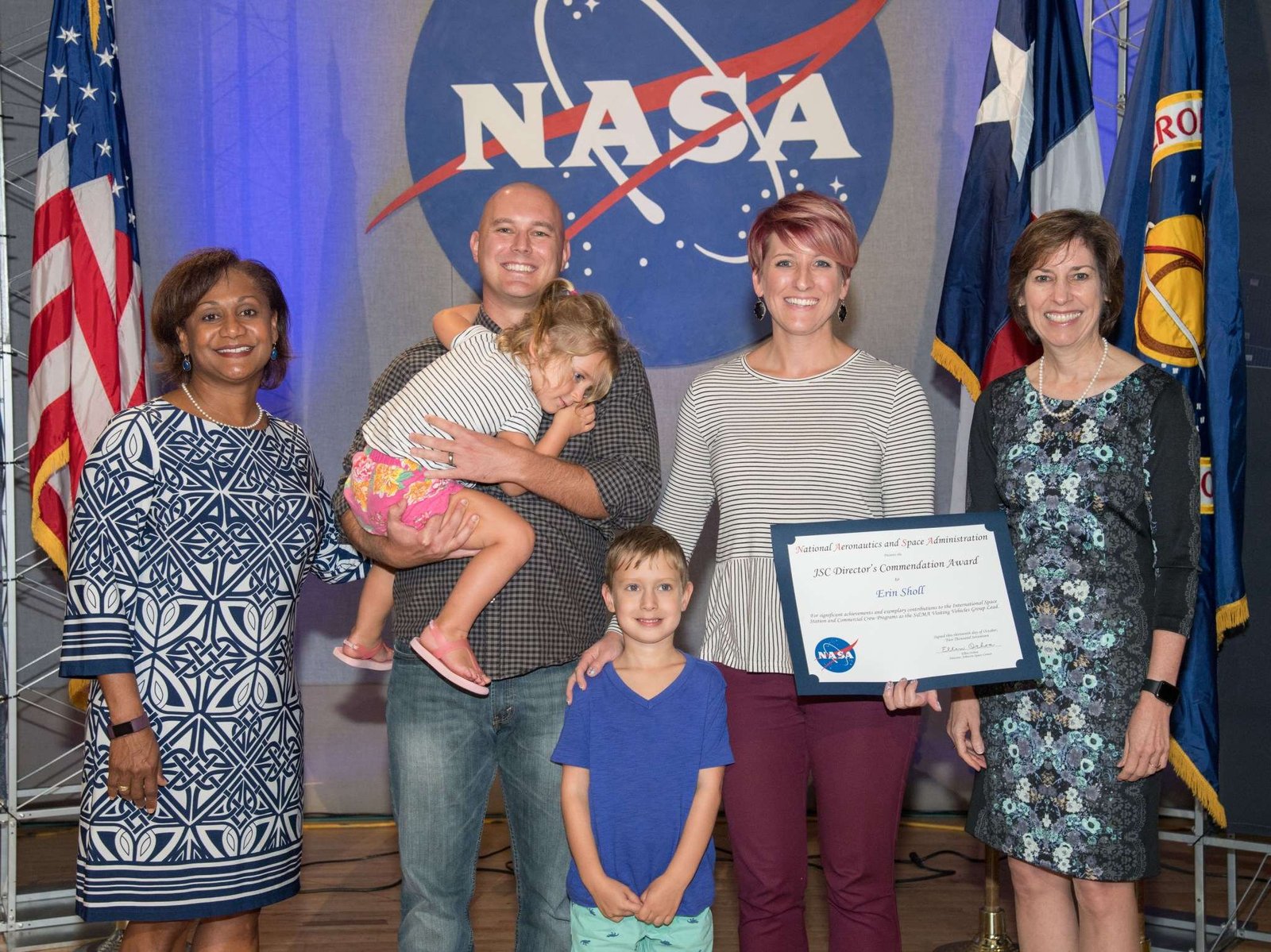 A woman receives a paper certificate on a stage with the American, Texas, and NASA flags. She stands with her husband, two children, and two other women in professional attire.