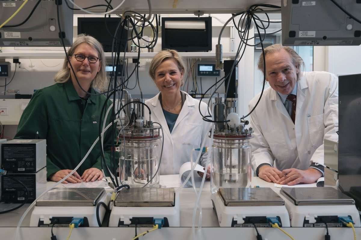 Two women and a man, all dressed in laboratory clothing, and in a laboratory setting, face the camera, smiling, and with documents arrayed before them on a workbench, while wires, weighing scales and other equipment is visible in the foreground and behind them