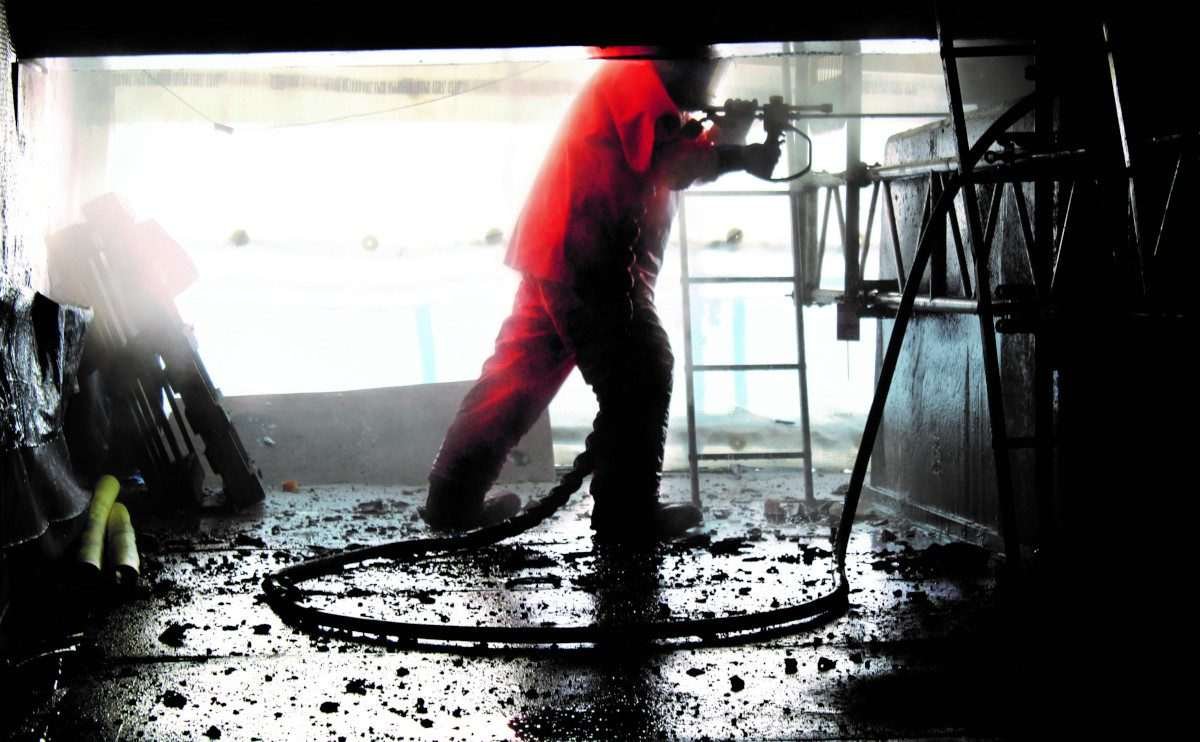 Torso and legs of man in protective clothing standing with one leg forward and performing some kind of operation that involves a hosepipe that trails along the ground