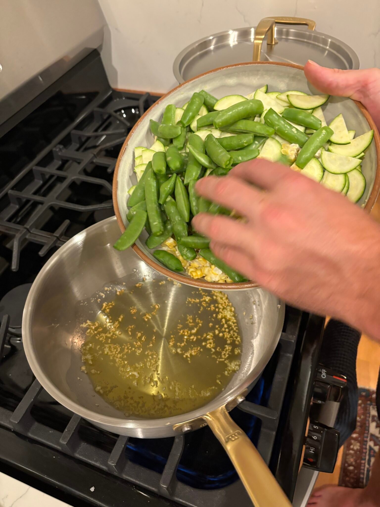 A person adds sugar snap peas and sliced zucchini from a bowl into a saucepan with sizzling garlic and oil on a stove.