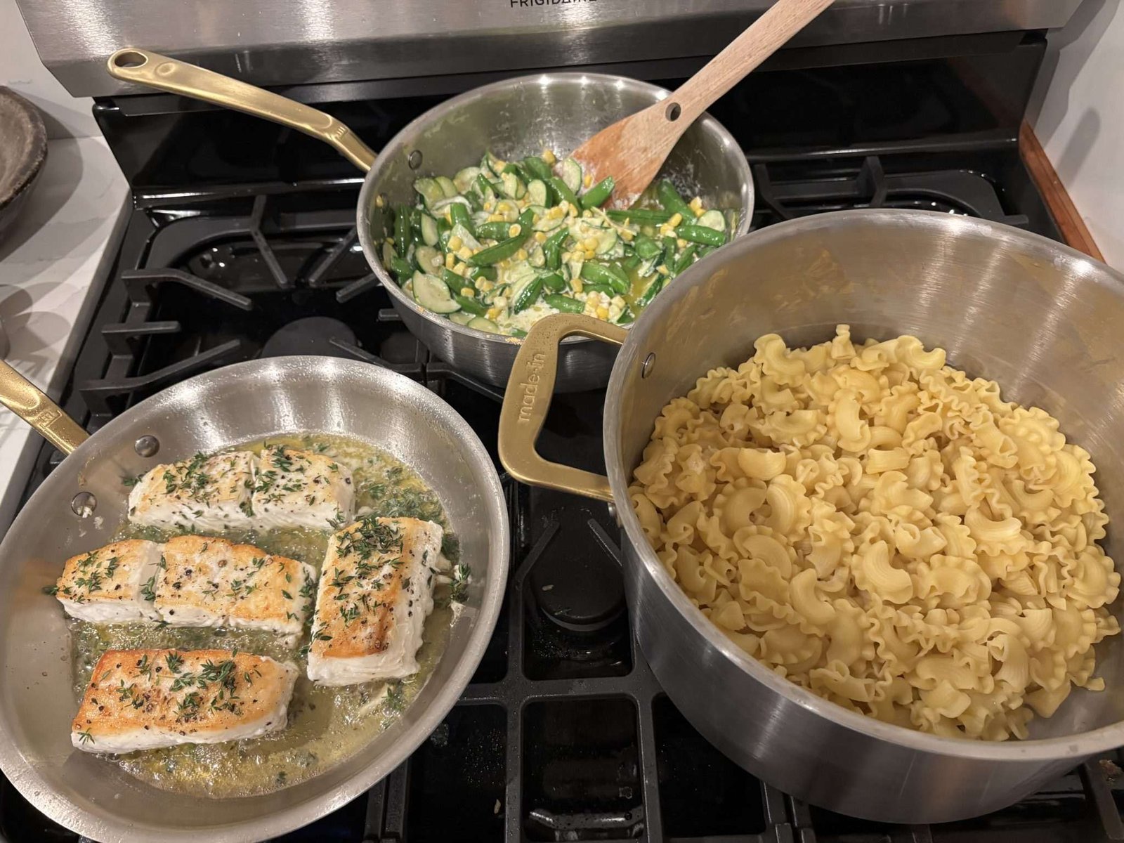 Three pans on a stovetop: one with cooked salmon fillets in herbs, one with sautéed zucchini and corn, and one with cooked pasta.