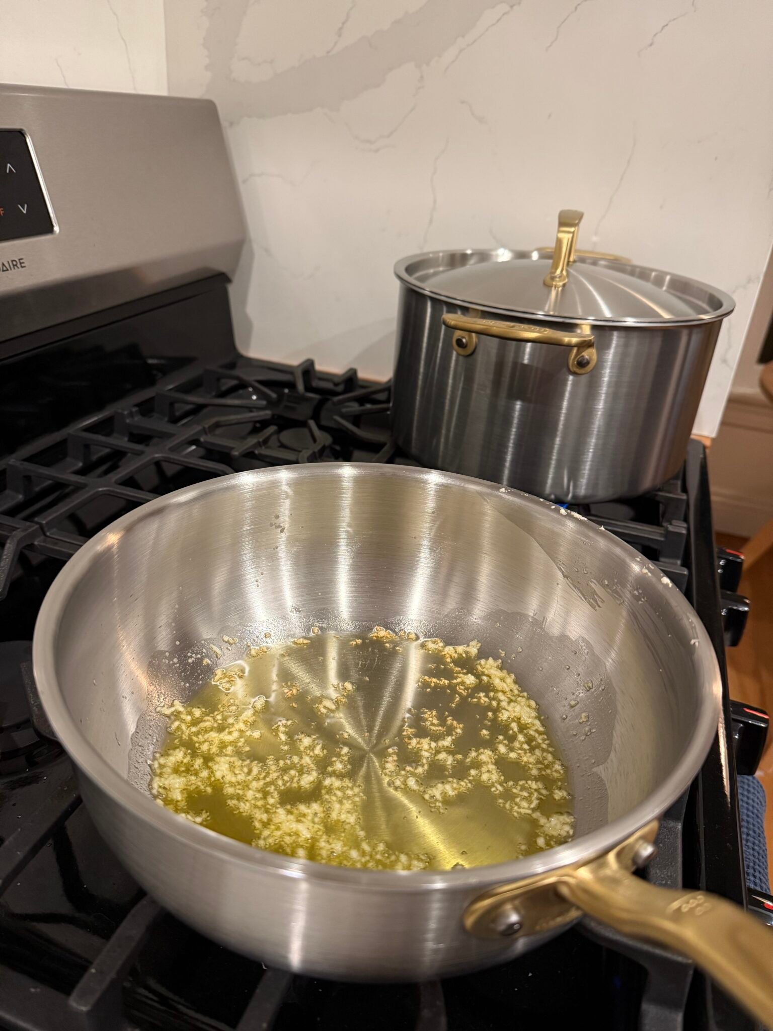 Minced garlic sautéing in oil in a stainless steel saucepan on a stove, with another lidded pot in the background.