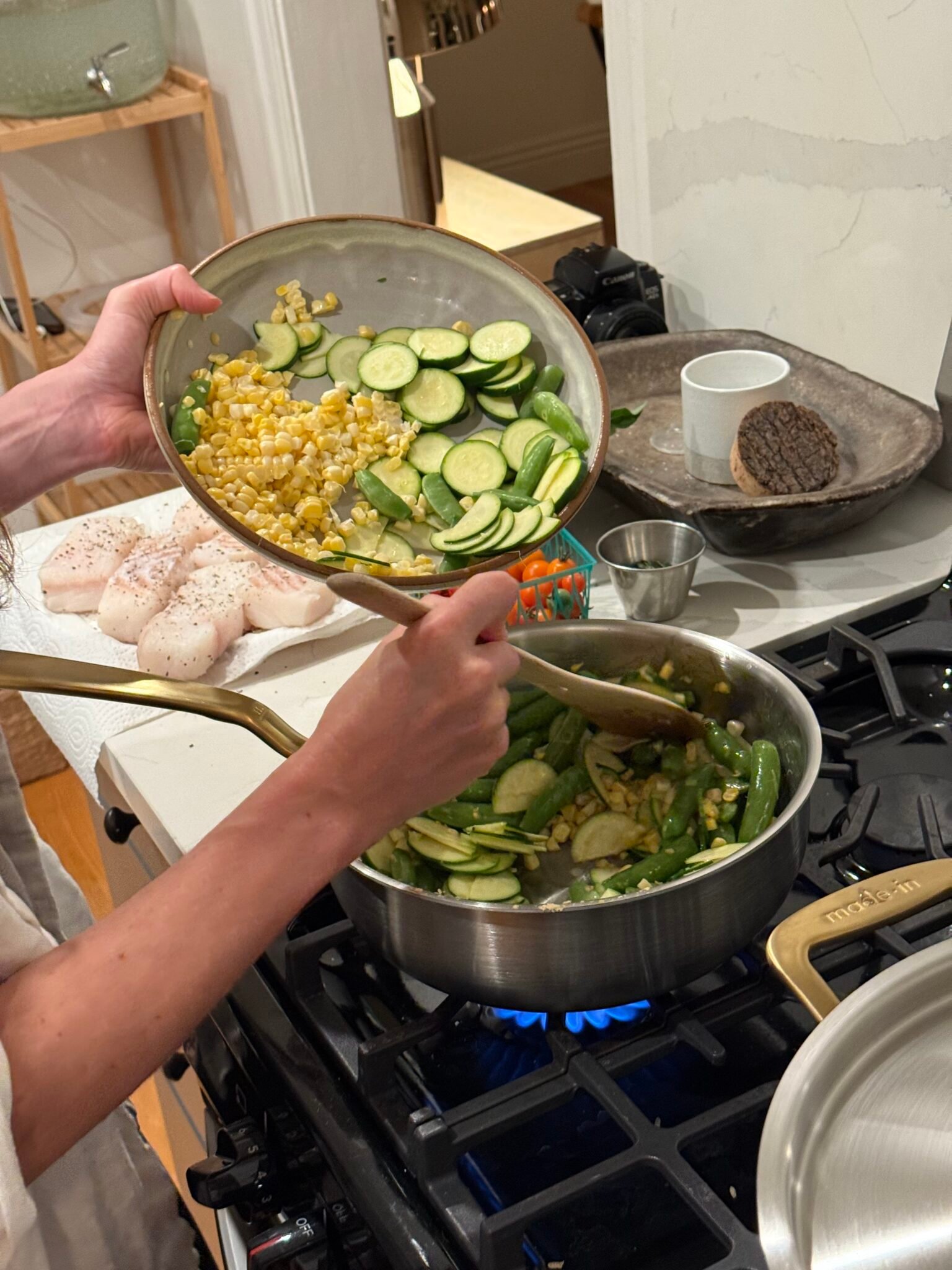 A person adds sliced zucchini, corn, and snap peas from a bowl into a pan on a stove. Raw fish fillets and ingredients are arranged nearby.