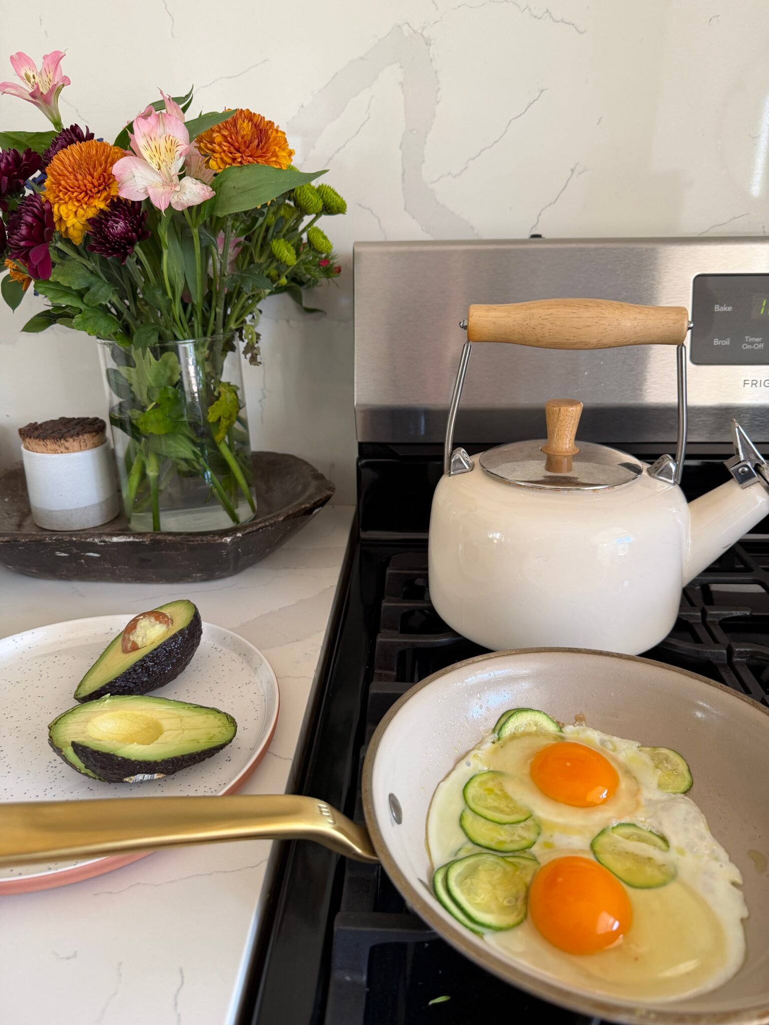 A frying pan with eggs and zucchini cooks on a stove next to a white kettle, with a plate of sliced avocado and a vase of flowers nearby.