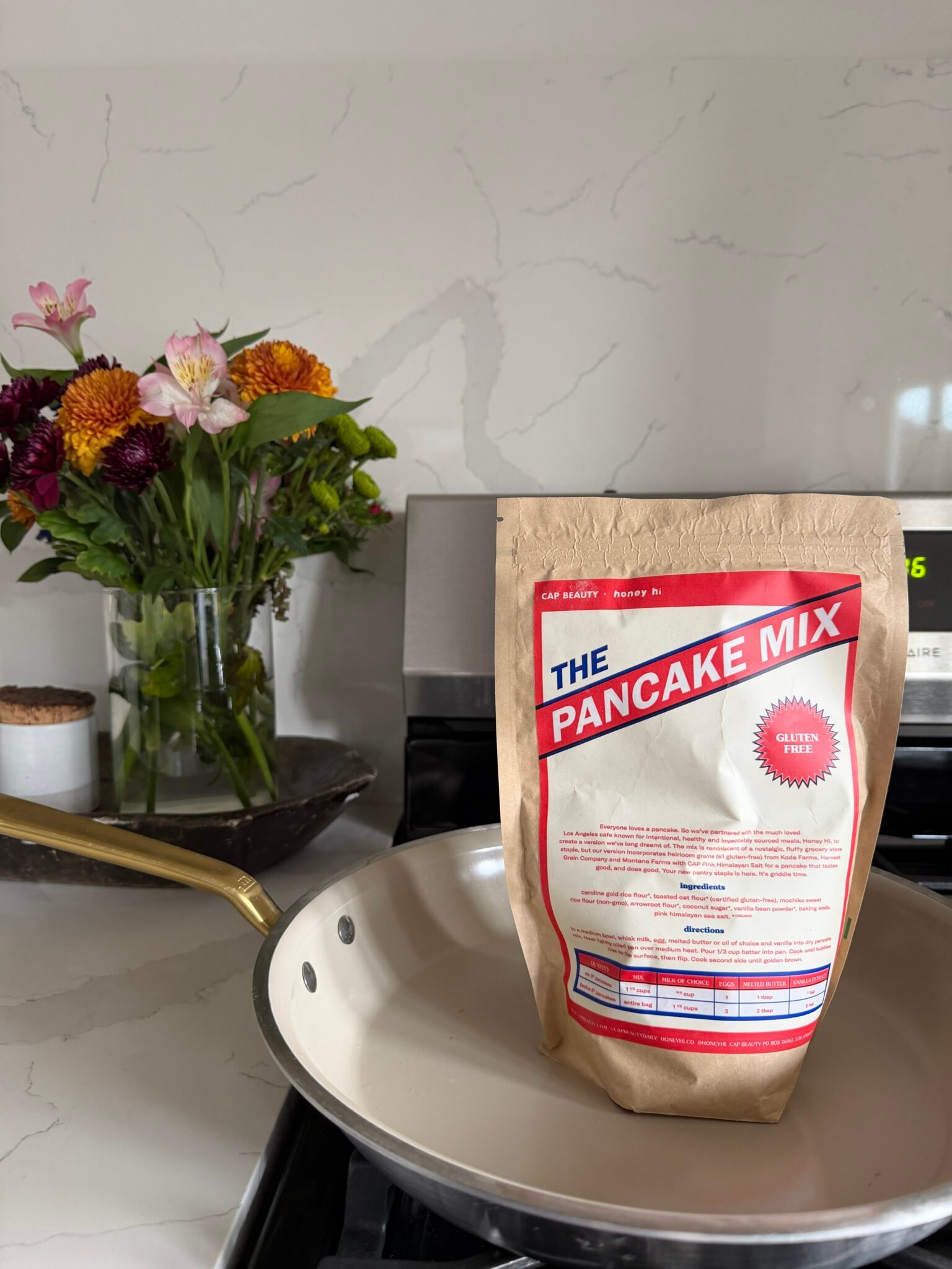 A bag labeled "The Pancake Mix" stands upright in a skillet on a stove, with a vase of flowers in the background.