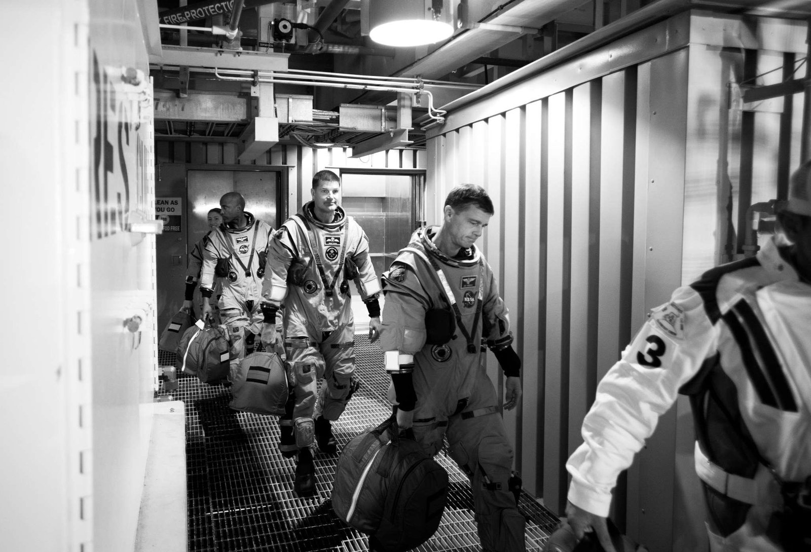 Four astronauts in spacesuits walk through a small corridor, following another person. The photo is in black and white.
