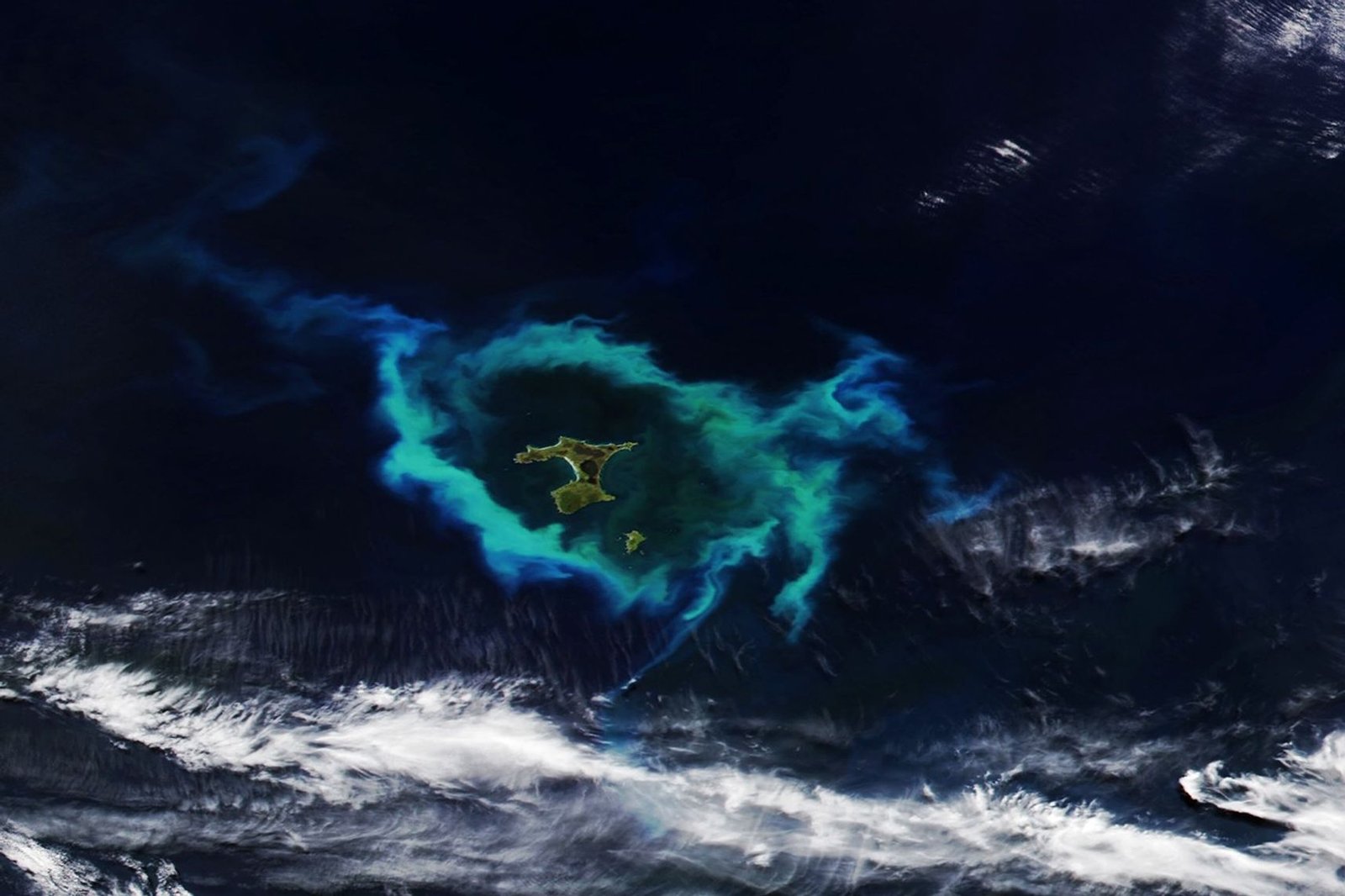 A phytoplankton bloom in the southern Pacific Ocean appears as an oval-shaped area of green and chalky blue water surrounding a group of small islands.
