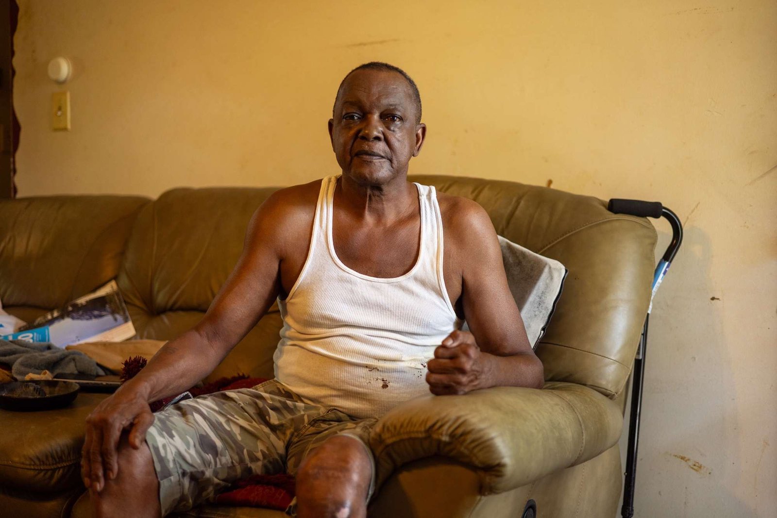 A man in a tank top sits on a brown couch with a cane nearby