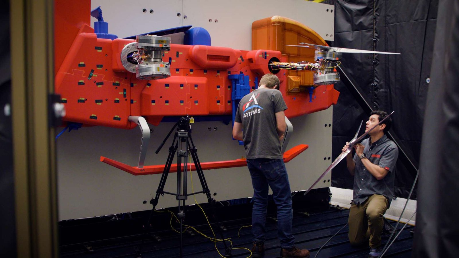 Two men in dark shirts work on a red car-sized rotorcraft protype in a testing chamber.