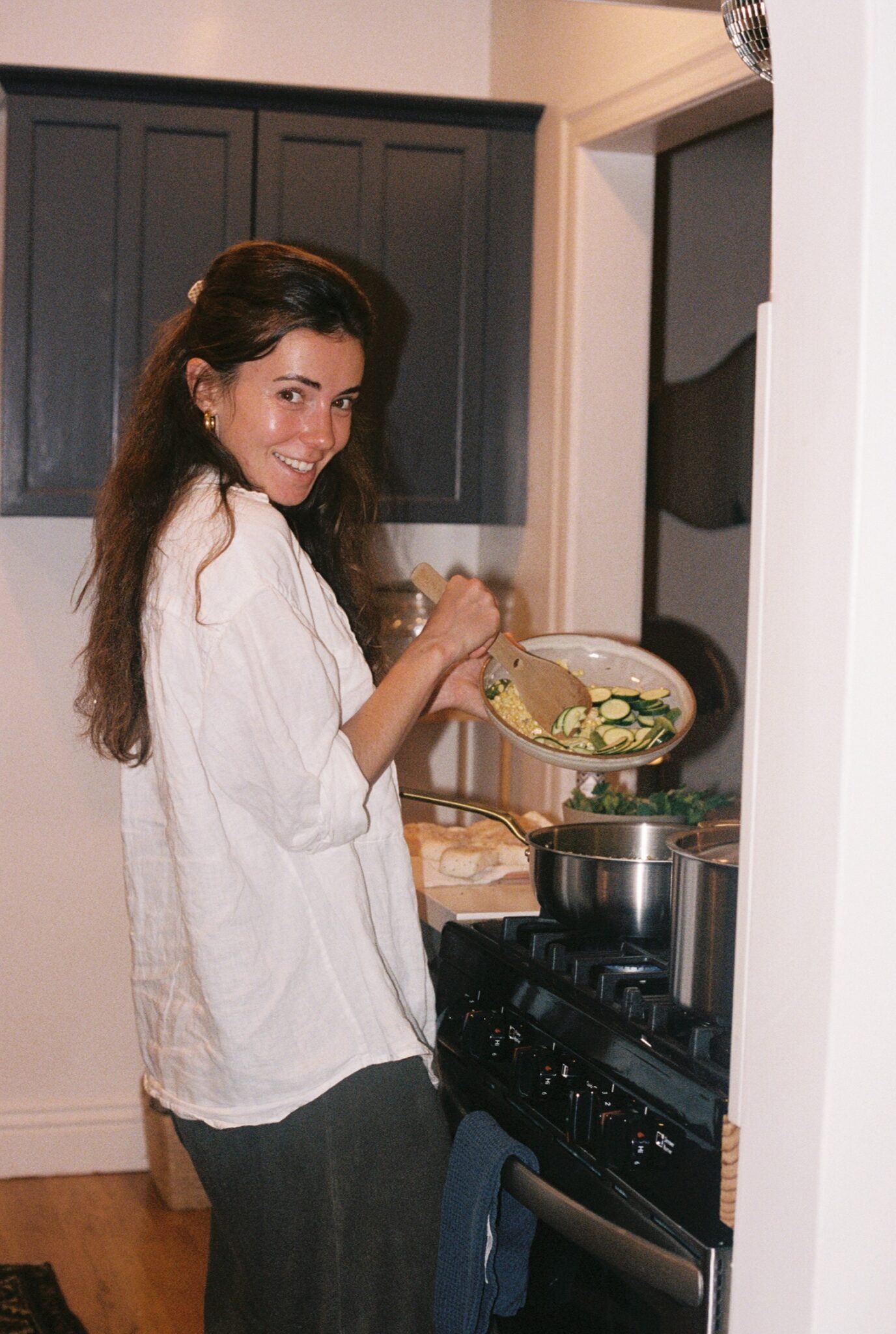 A woman stands at a stove, stirring vegetables in a pan with a wooden spoon, while smiling at the camera in a home kitchen.