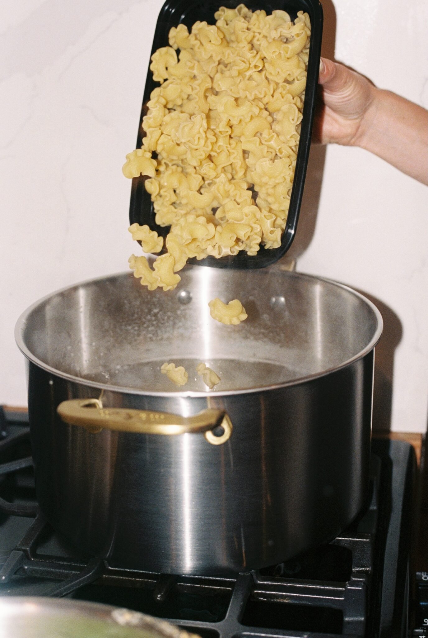 A hand pours dry pasta from a black container into a pot of boiling water on a stove.