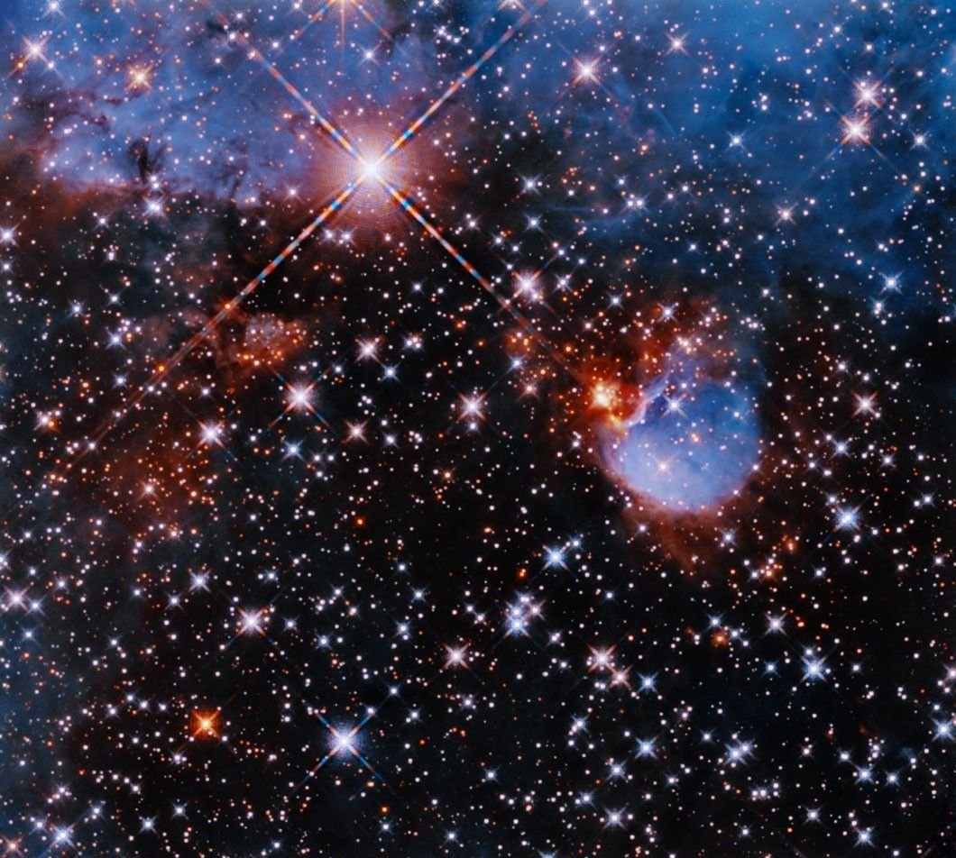 Within a field of glittering multicolored stars, bluish clouds of gas edged in glowing red cluster at the top of the image and in a bubble-shaped clump to the mid-right. A single shining star throws diffraction spikes across the upper left of the image.