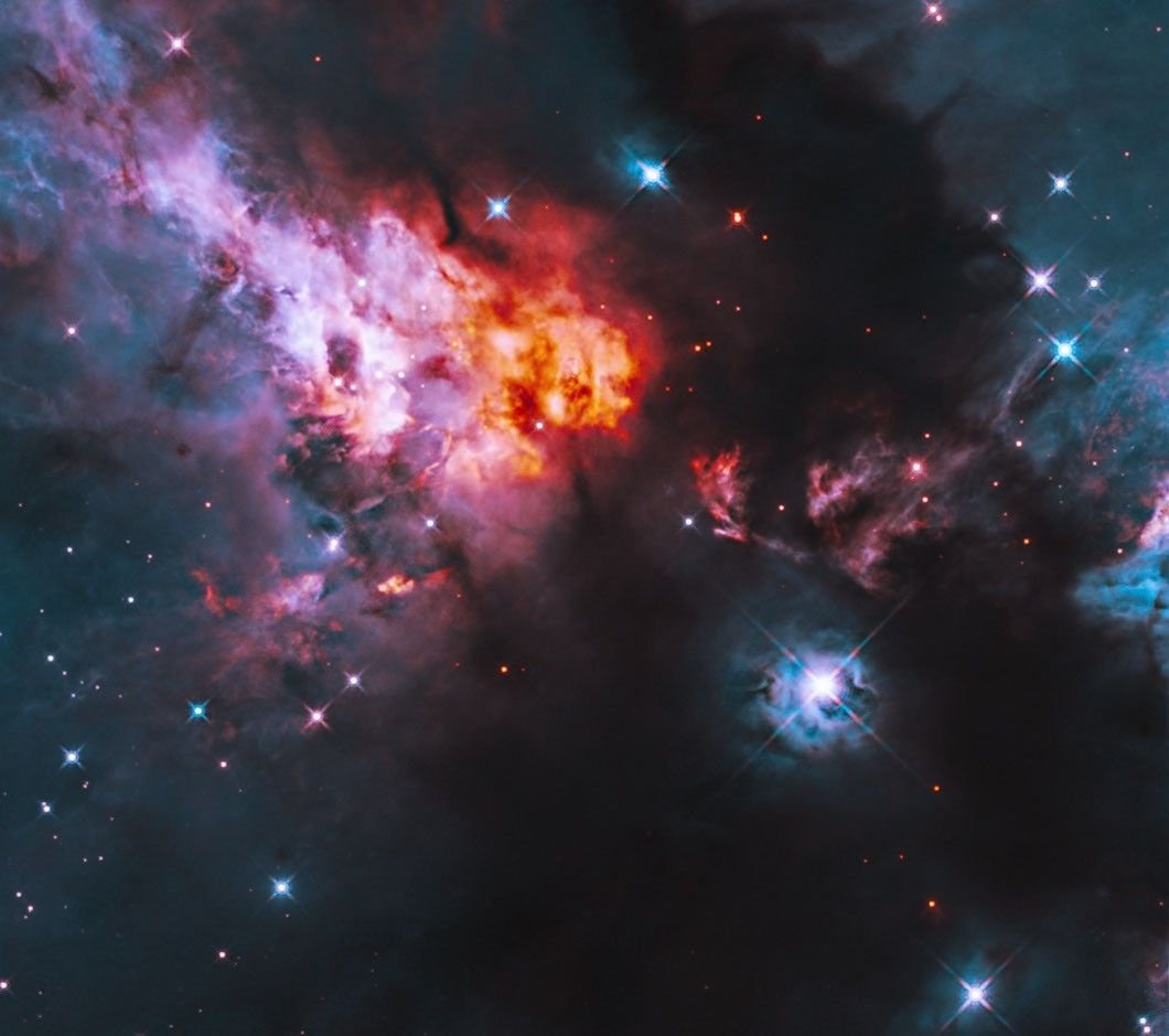 Shining blue stars are sprinkled throughout glowing clouds of orange, pink and bluish gas, alongside dark clouds of dust. A particularly bright star shines against the inky dark dust of the lower right quadrant.