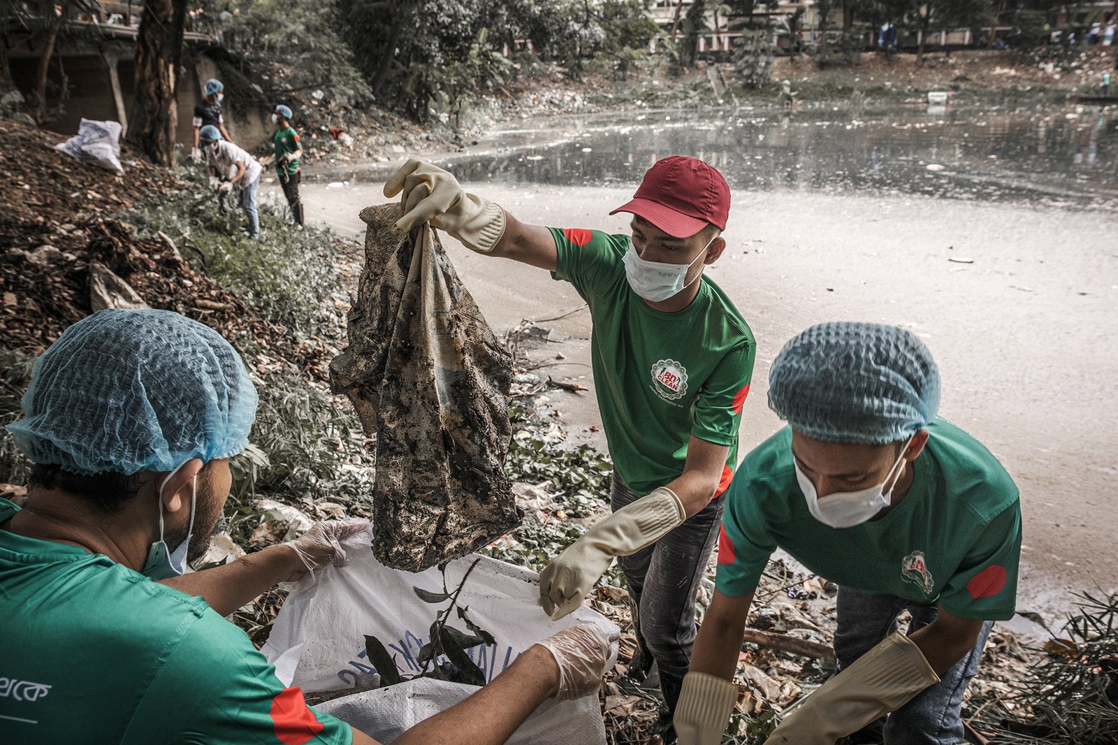 A group of people in matching teal t-shirts pick up trash from near a bod of water