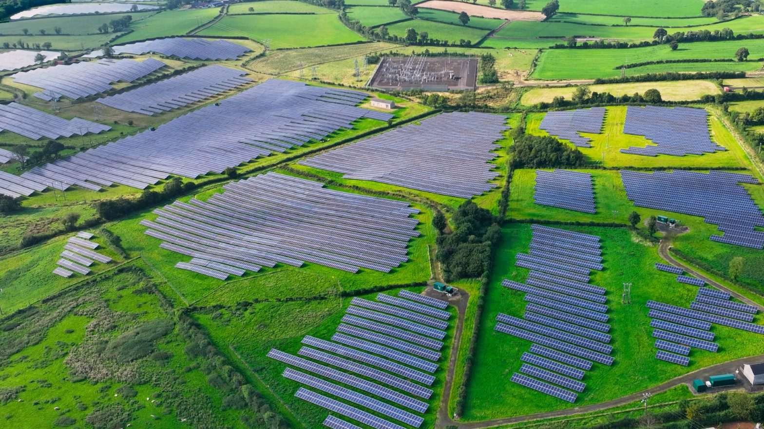 Aerial view of green fields in which long, rectilinear grey structures are arrayed - presumably solar panels