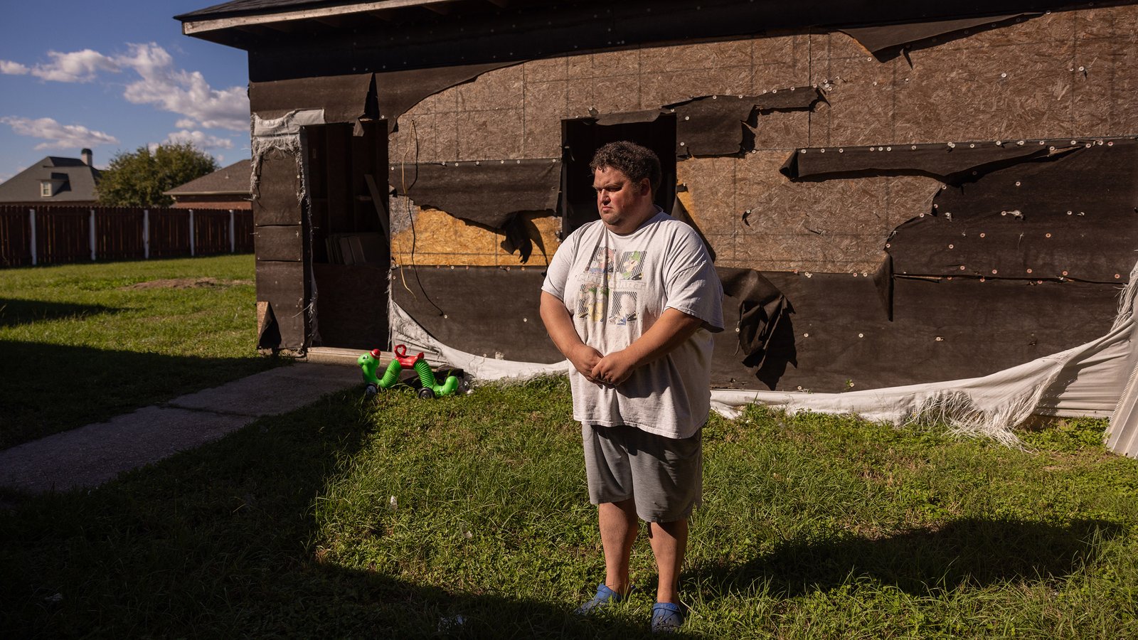 A middle-aged man in a mariokart t-shirt stands in front of a house with a severely damaged facade covered in torn construction tarp and mesh. Near the house, a small green plastic worm toy stands propped up next to a sidewalk