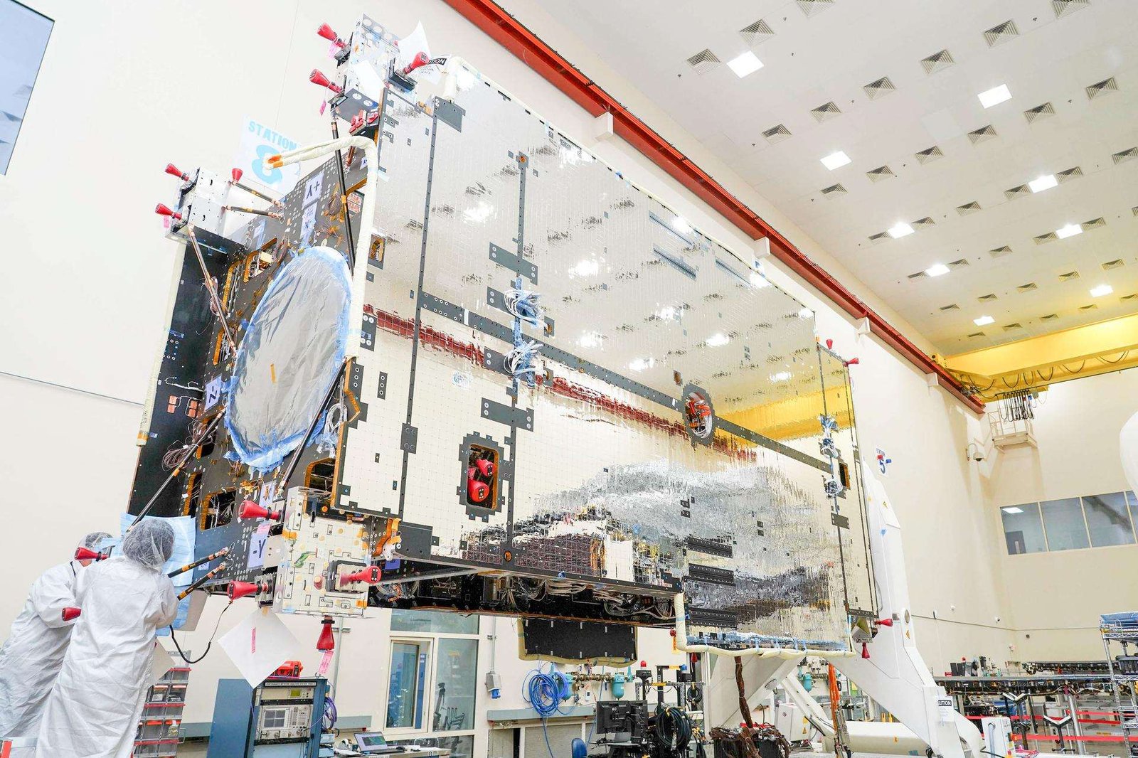 The primary structure of Gateway’s Power and Propulsion Element stands inside a high-bay cleanroom at Lanteris Space Systems in Palo Alto, California. The large rectangular structure is covered in reflective silver-colored panels. Two technicians in white cleanroom suits work near the base of the structure. 