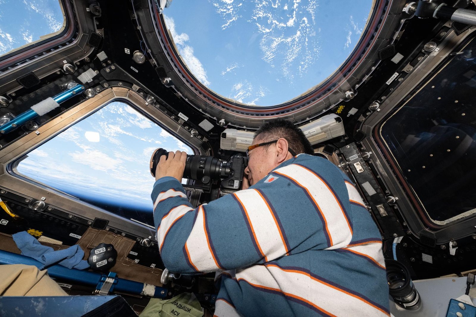 JAXA (Japan Aerospace Exploration Agency) astronaut Kimiya Yui, wearing a blue-and-white striped shirt, looksthrough a camera inside of the space station’s cupola. The blue Earth, speckled with clouds, can be seen through a circular window above and trapezoid-shaped windows in front of him.