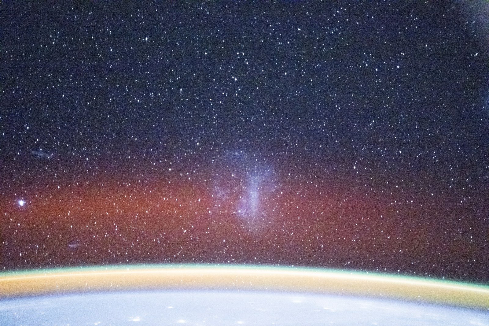 The blue curvature of the Earth’s atmosphere is visible with yellow, green, and red layers of airglow above it. In the center, just above Earth and peeking through the red haze, a blue collection of stars is visible.