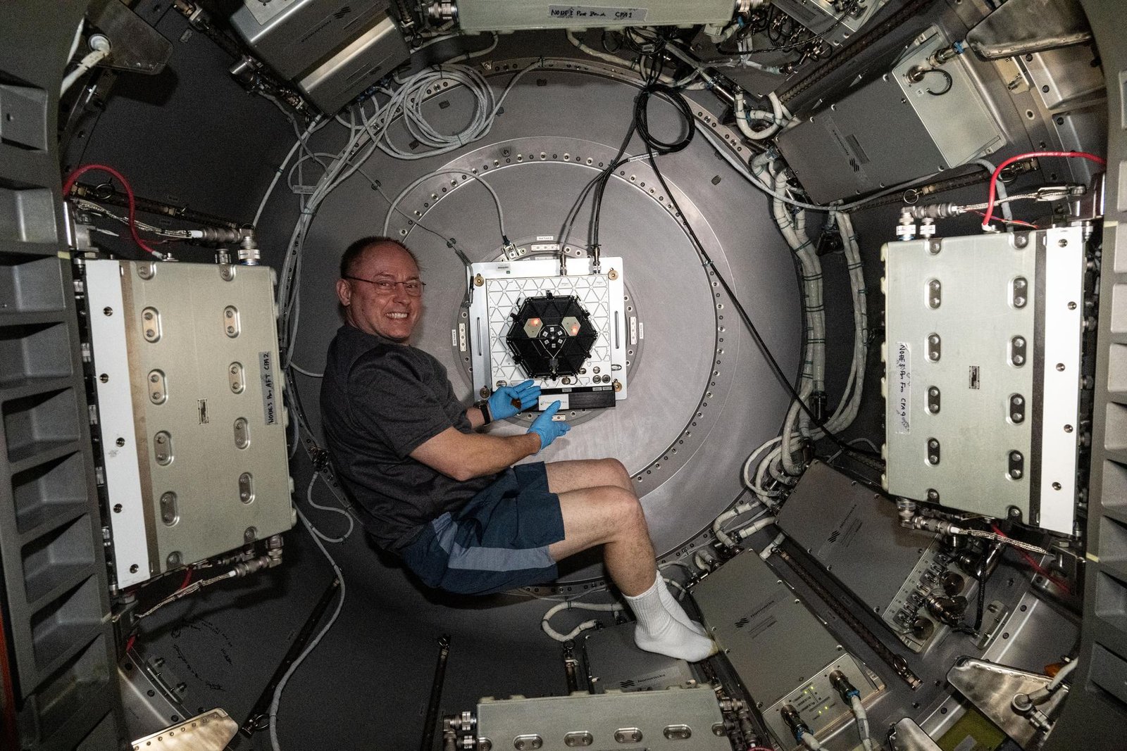 NASA astronaut Mike Fincke floats in a cylindrical airlock. He wears blue latex gloves and gestures toward a silver box with a black, hexagonal shape in the center. He is surrounded by wires and various panels along the space station’s walls.