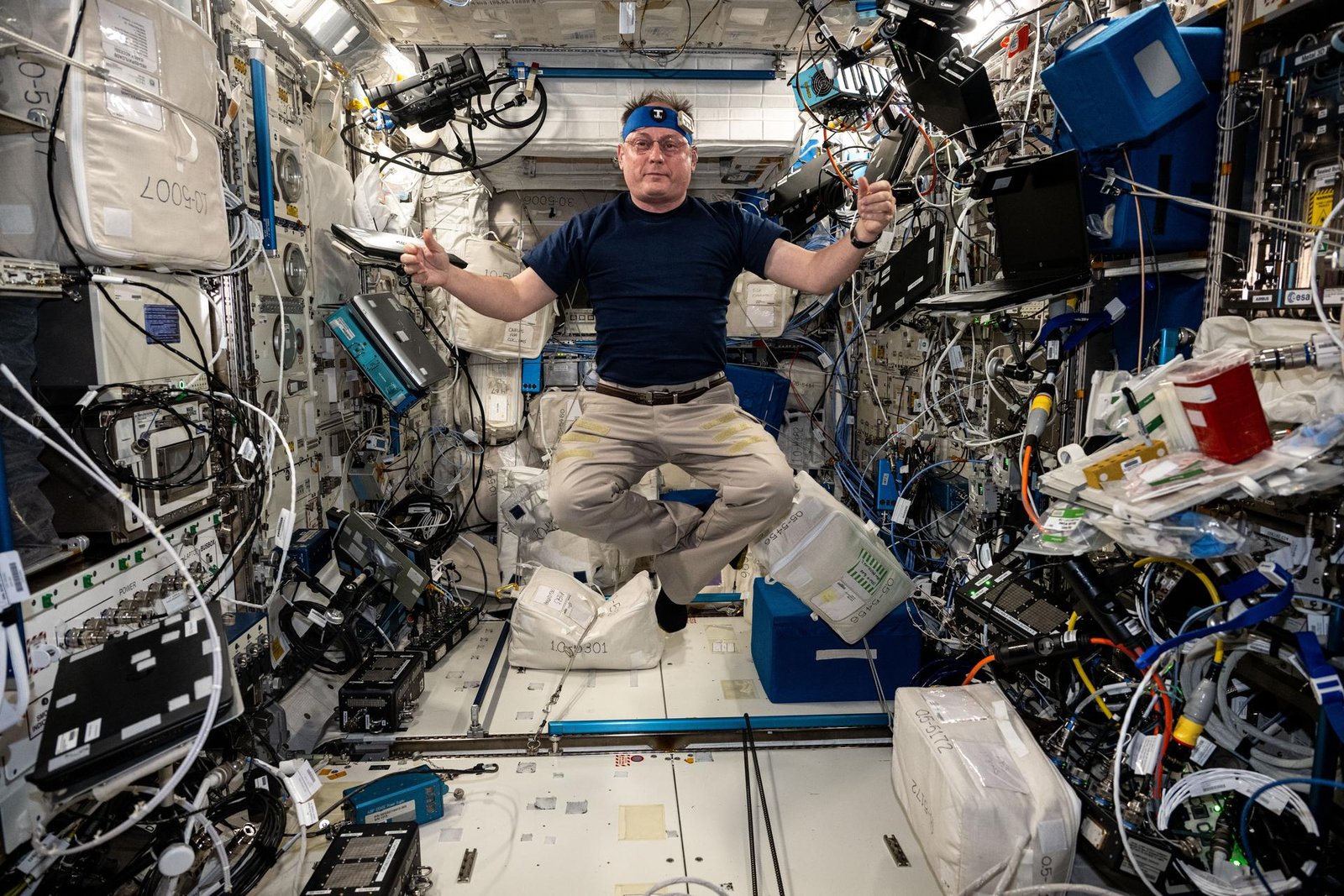 NASA astronaut Mike Fincke, wearing a black shirt and khaki pants, floats in the space station with his arms up and legs crossed. He wears the blue T-Mini headband. The surrounding walls of the space station are lined with equipment and wires.