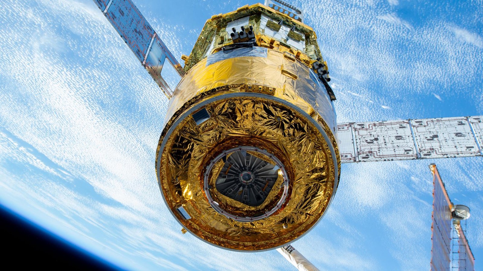 The cylinder-shaped HTV-X1 spacecraft is shown gripped by the Canadarm2. The surface of the vehicle is reflective gold, and a silver-colored solar array panel juts from either side. The Earth fills the background, with white streaky clouds covering much of the blue ocean.