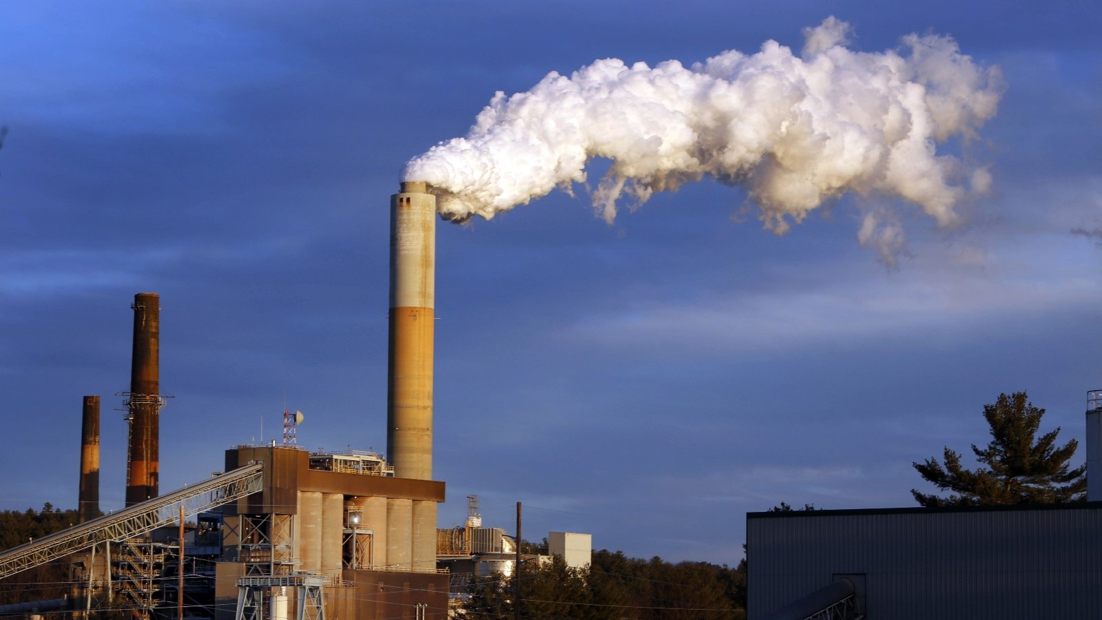 A power plant with smoke stacks belch smoke against an evening sky