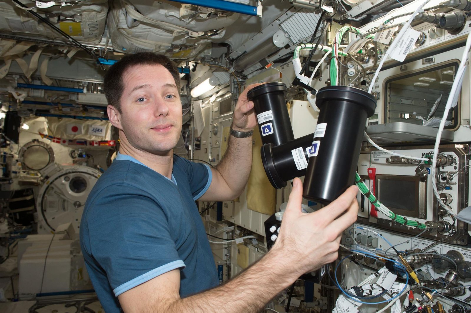 European Space Agency astronaut Thomas Pesquet works inside the International Space Station, holding two black cylindrical experiment containers as part of the Protein Crystal Growth-5 study. Behind him, computer displays are mounted to the station’s wall, along with many other instruments, cables, and silver knobs.