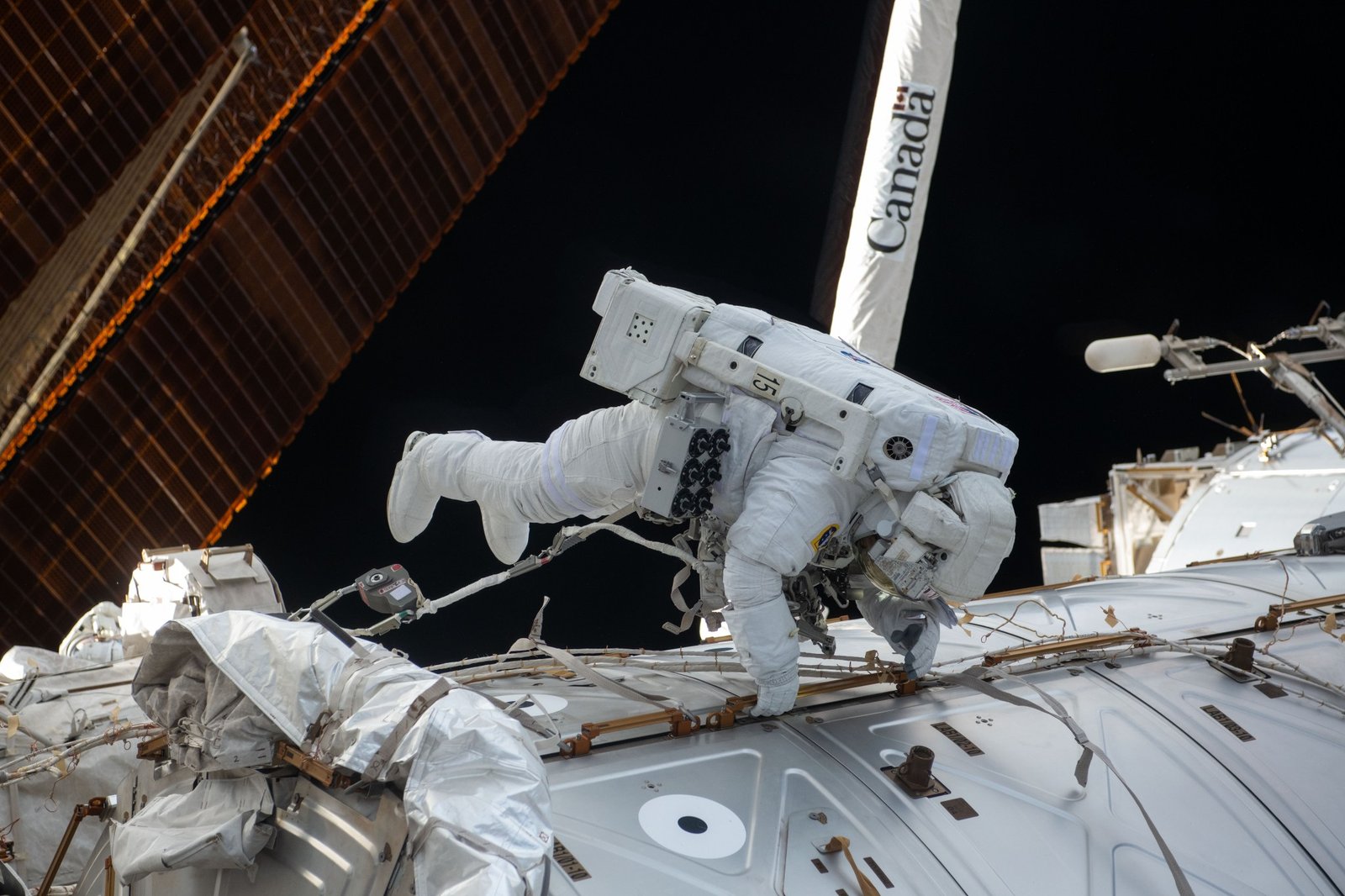 NASA astronaut Butch Wilmore wears a white spacesuit while performing a spacewalk outside the International Space Station. Wilmore is positioned horizontally against the station’s exterior. In the background, the black of space contrasts with the station’s copper-colored solar arrays and the white robotic Canadarm, which has "Canada" printed vertically in black letters.
