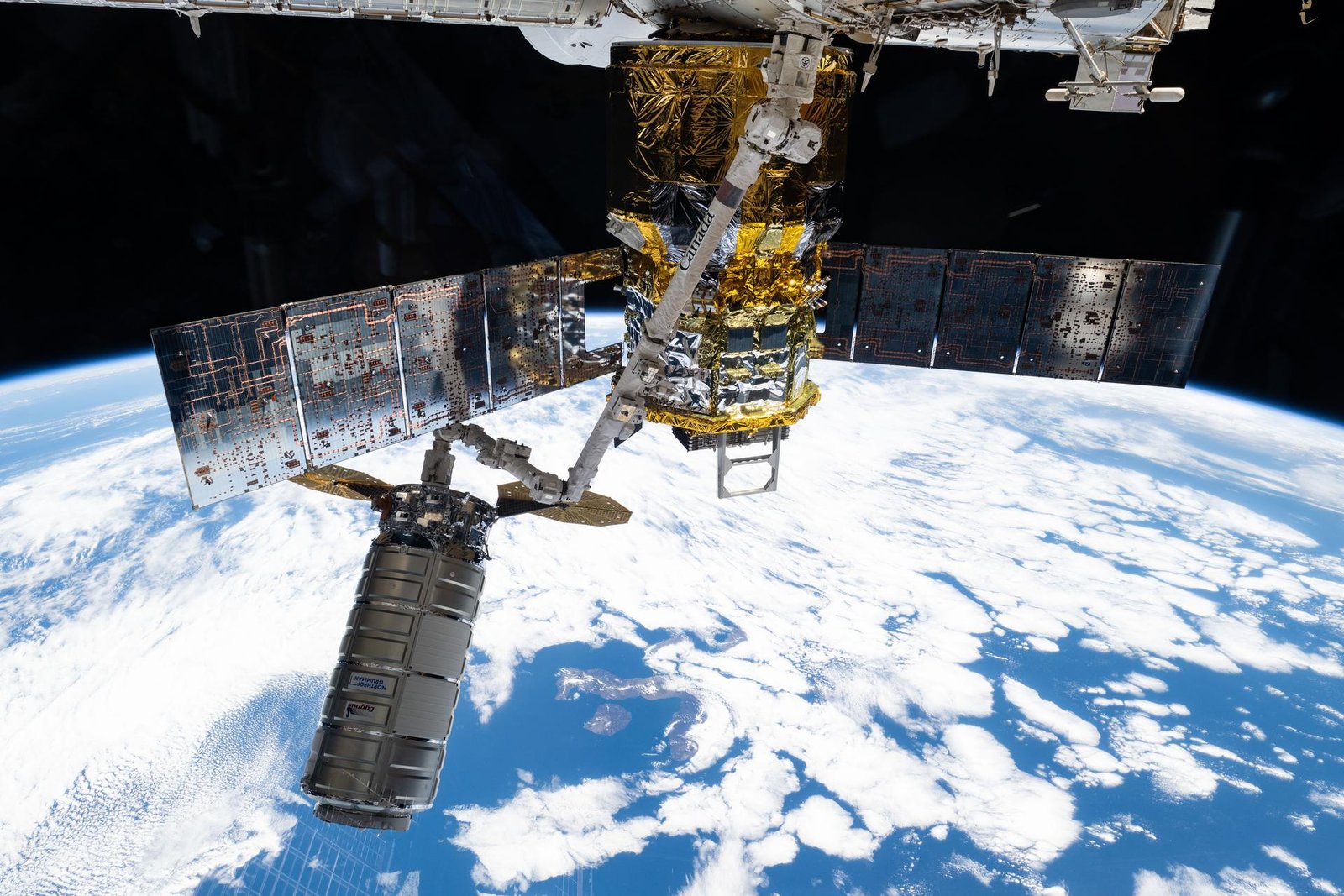 A shot from the International Space Station showing a long, white robotic arm maneuvering the cylindrical silver Cygnus XL cargo spacecraft. Earth’s blue oceans and white clouds are visible in the background. In the foreground, the HTVX-1 vehicle docked to the space station, covered in reflective gold foil with its silver solar panels visible.