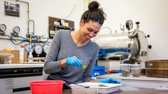 A woman, smiling, works at a laboratory bench, appearing to pour material from a small tub held in one hand onto a white tray or similar receptacle