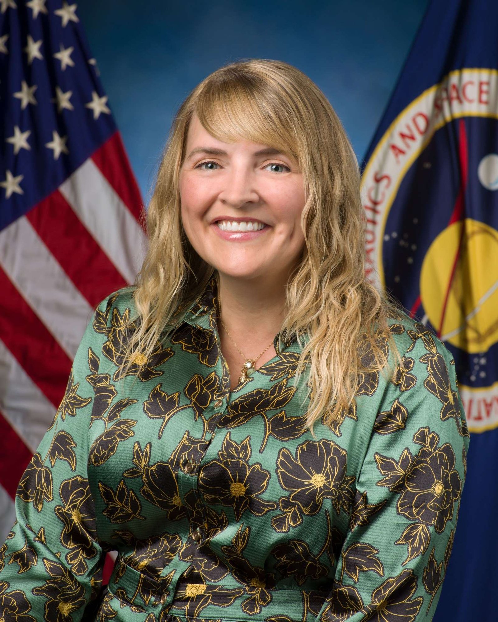 A woman wears a colorful blouse and poses in front of a blue background and a NASA flag (right) and U.S. flag (left).