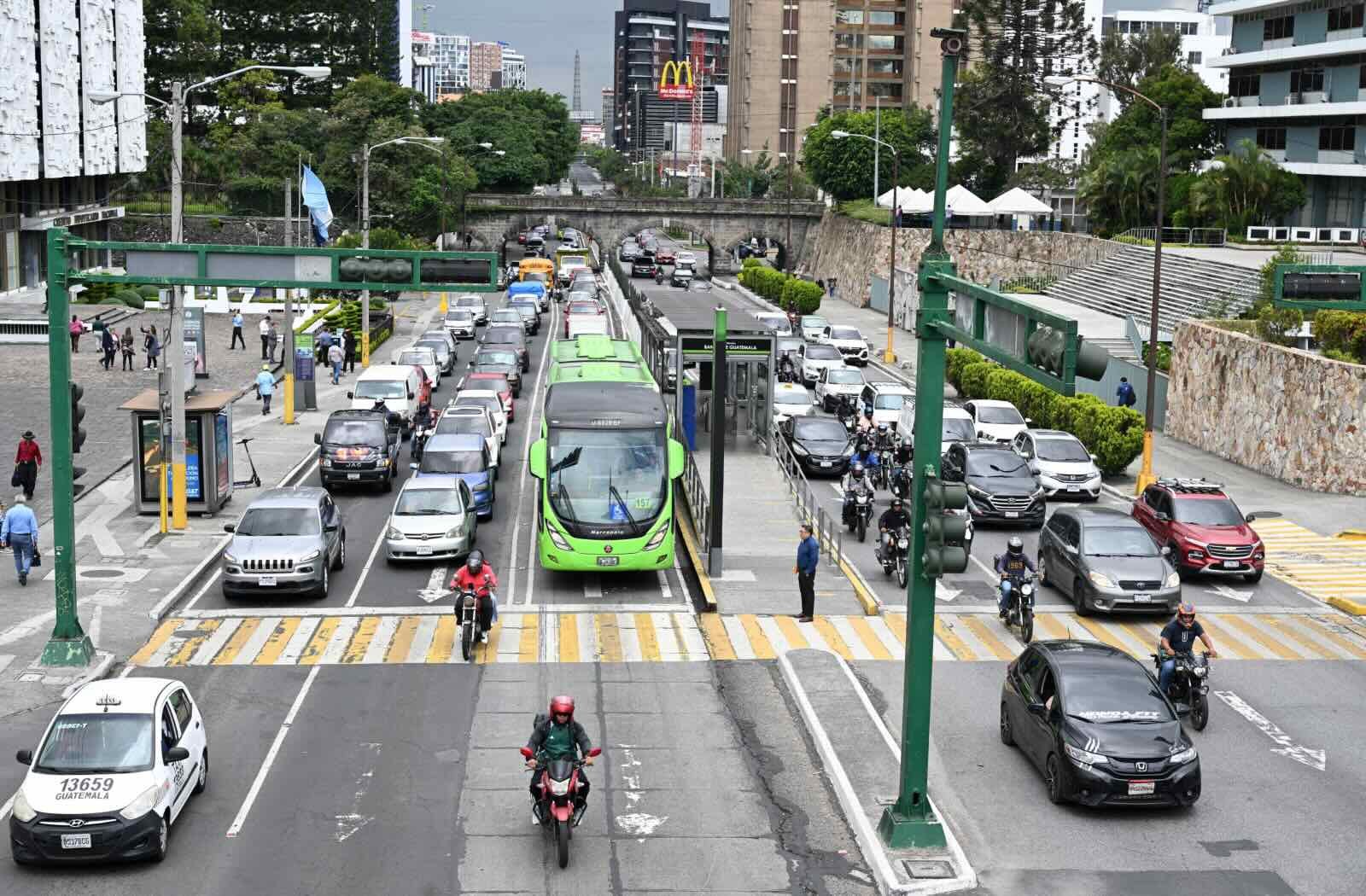 A photo shows a green bus, motorcycles, and cars stopped at a traffic light in a bustling downtown area