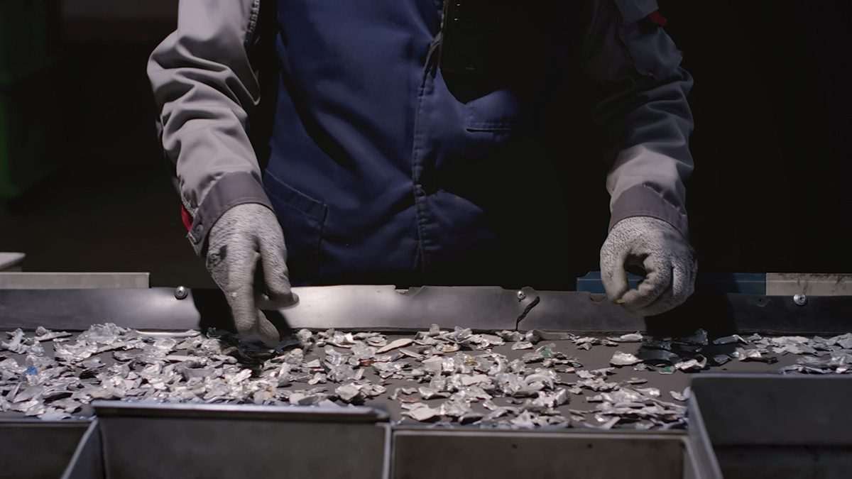 Metal fragments on a conveyor belt in a darkened space, with a pair of grey-gloved, overalls-wearing human arms in view at top of the image, picking up pieces of metal