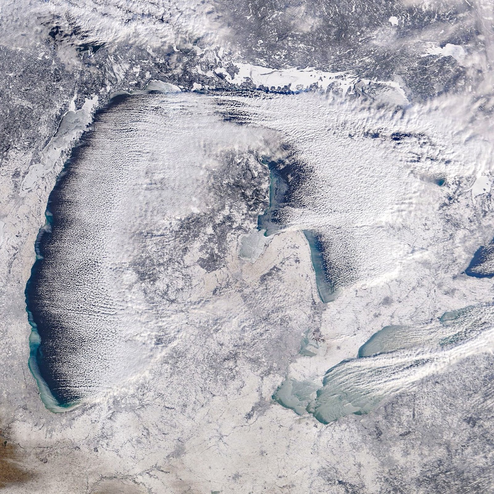 A satellite view of the Great Lakes shows a winter landscape, with snow-covered land, ice forming on parts of the lakes, and clouds trialing over open water.