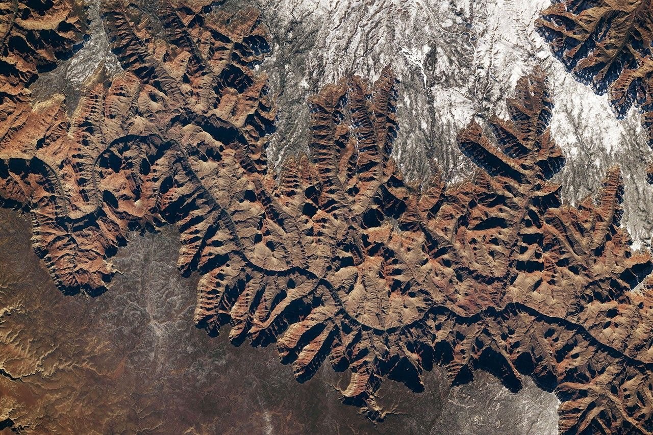 A portion of the Grand Canyon, with the curving Colorado River at its bottom, runs across the width of this downward-looking photo. Snow covers some of the plateau on both sides of the canyon, with more appearing on the North Rim.