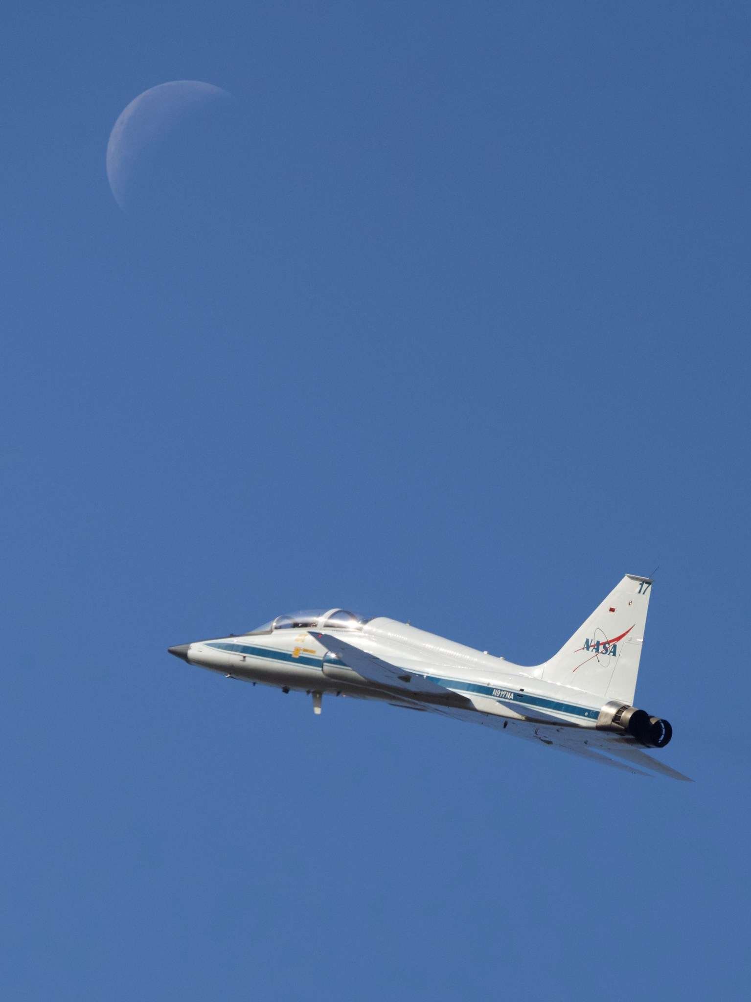 A white plane rises upward to the left against a blue sky. At top left, the waning crescent moon is faintly visible. The plane has a blue stripe low on its body and a version of the NASA insignia on the rudder.