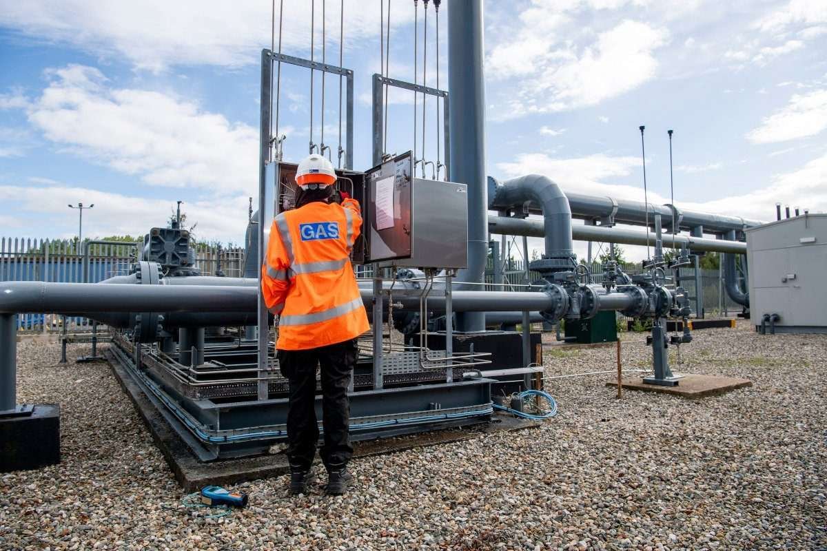 A gas engineer in orange luminescent vest and hard hat, standing with back to the camera, is at work on a control panel in a small cuboidal metal enclosure within a gas network outdoor site, with extensive pipework and other objects visible