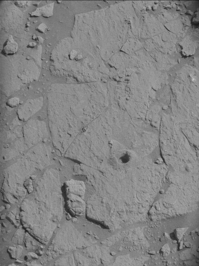 An overhead, grayscale photo of the Martian surface shows a uniformly medium gray, flat, rocky area, where a few wide, shallow cracks on the surface are filled with small rocks. A drill hole, fully shadowed so you can’t see its depth, is in the ground near the center of the frame.