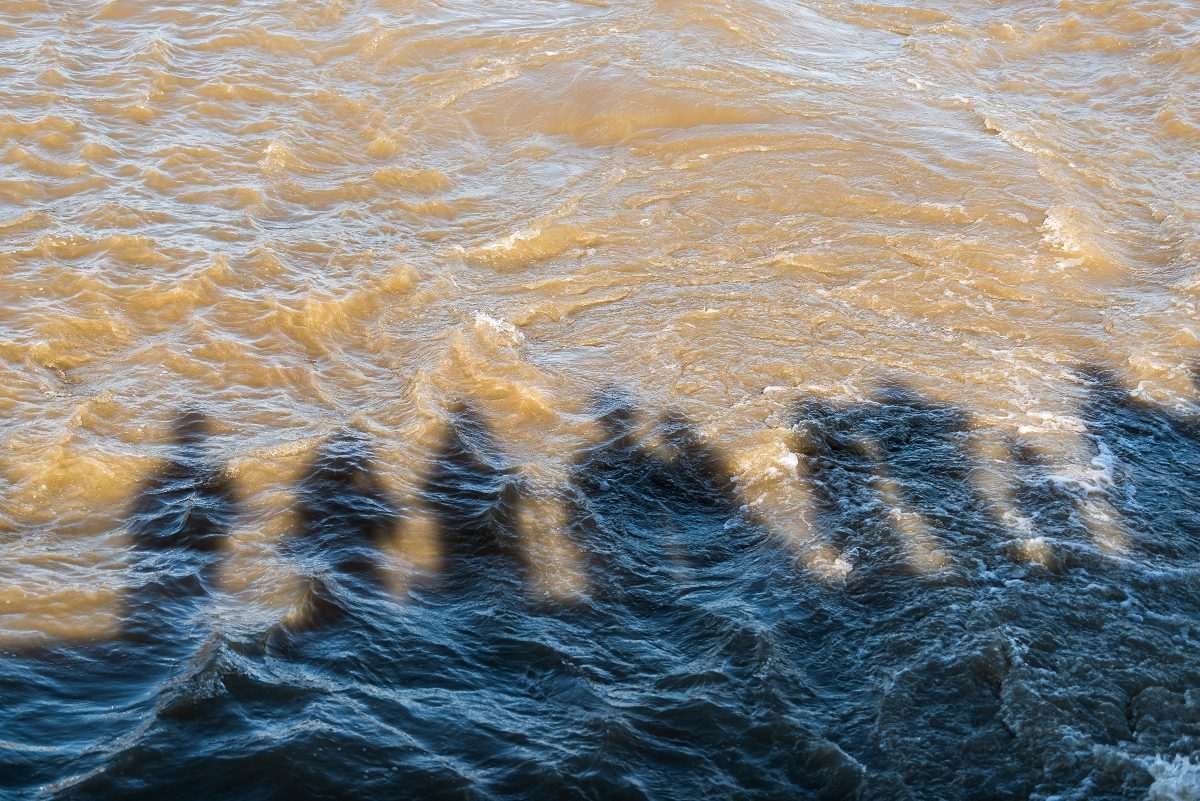 Shadow cast by a row of people standing (on a bridge or river bank perhaps) onto a brown or muddy river surface