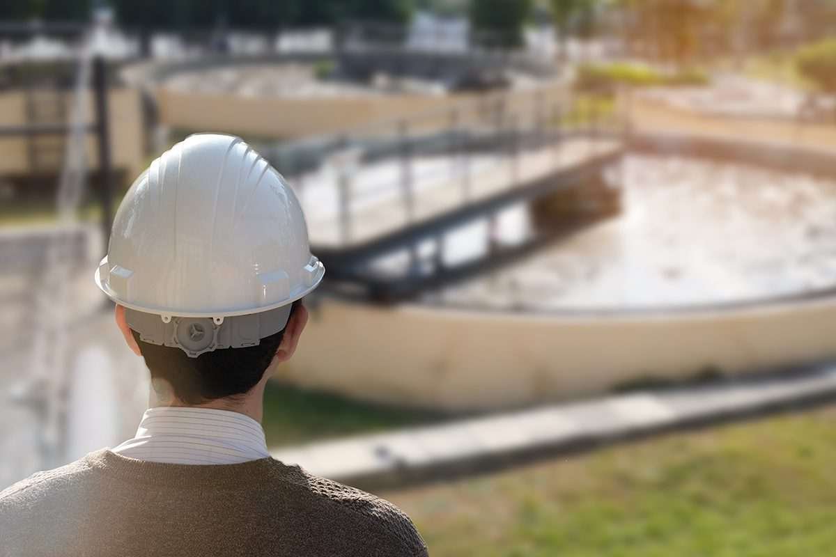 View from just behind the head of an engineer wearing a white hard hat, looking towards a landscape that includes a circular water treatment tank