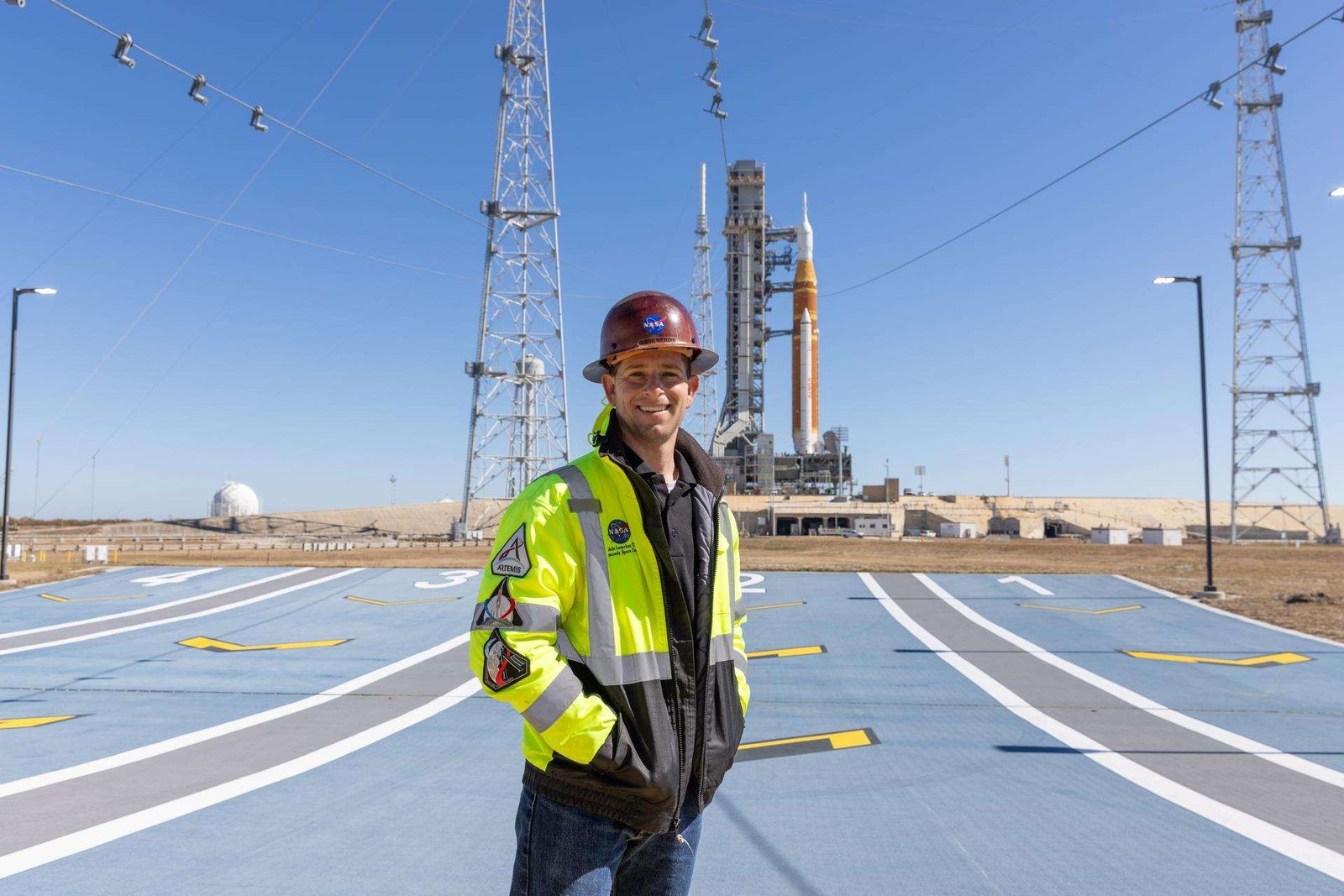 Image shows Jesse Berdis standing standing at the pad of Launch Complex 39B at NASA's Kennedy Space Center in Florida. Behind him are clear blue skies and NASA's SLS (Space Launch System) rocket and Orion spacecraft for the Artemis II mission. Photo credit: Kim Shiflett