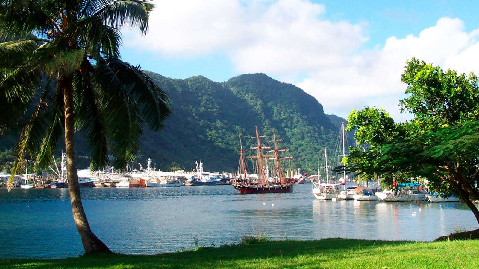 A sailing ship is seen in a harbor surrounded by lush palm trees and mountains on a sunny, idyllic day